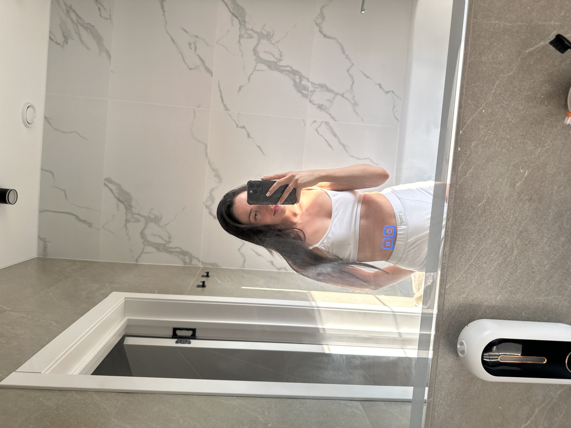 In a modern bathroom with marble-patterned tiles, a young woman with dark hair stands in front of a large mirror, taking a selfie with her smartphone. She is wearing white undergarments, a bralette and shorts. The mirror reflects the bathroom, including a doorway, a bathtub, and a soap dispenser mounted on the wall. There are two illuminated icons on the mirror in front of the woman, one resembling wavy lines and the other a power button. The location is identified as Эстачи, Russia, suggesting this is a residential bathroom.