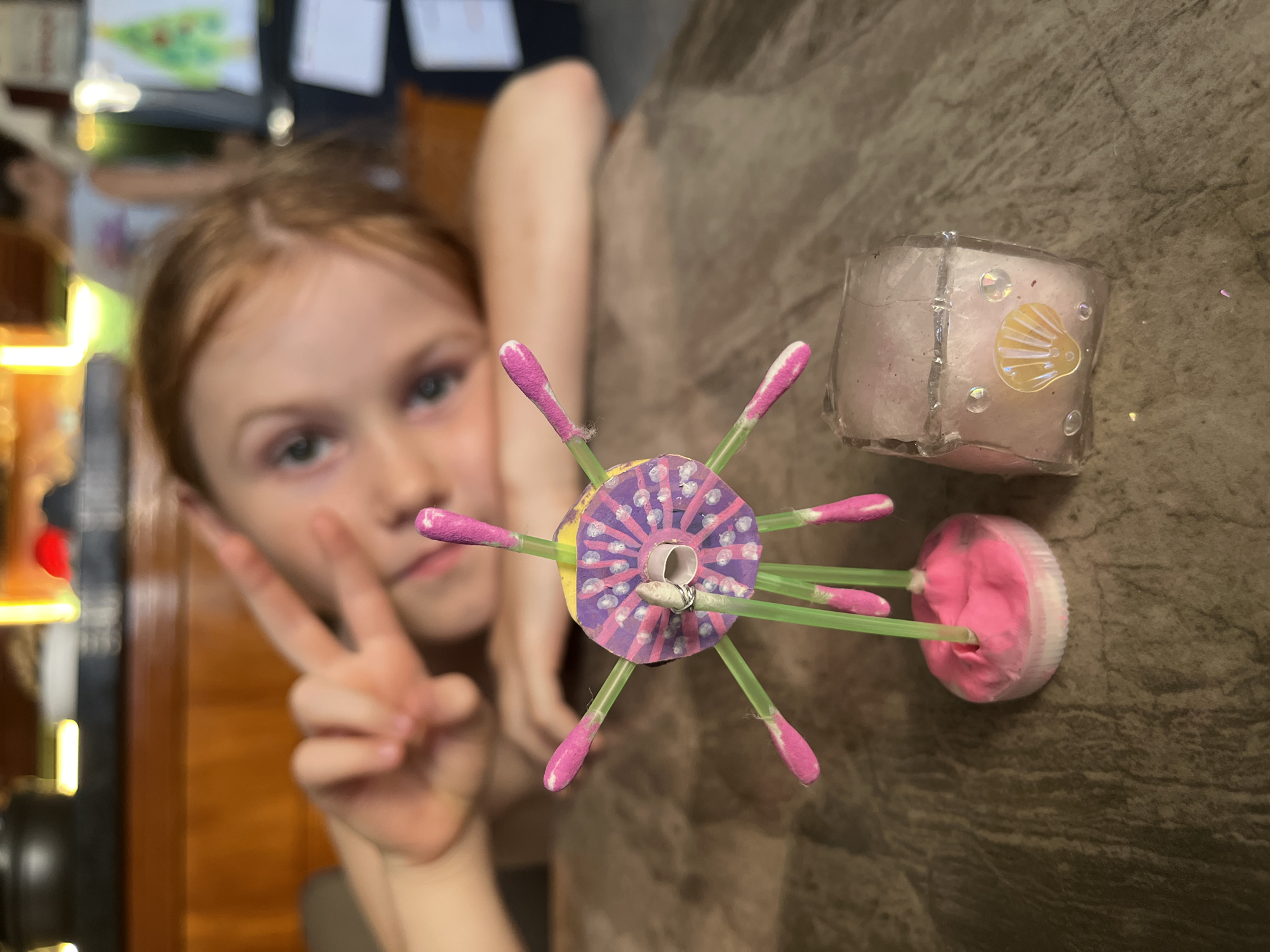 A young girl with reddish-blonde hair gives a peace sign, with a crafts project visible in the foreground. The craft appears to be made of a decorated purple circle with green straws sticking out and pink cotton swabs on the ends. Beside it is a translucent cube object with a seashell embellishment. The setting looks like an interior space with a wood-look surface. There is no information to identify this as Bac Nha Trang Ward, Vietnam.