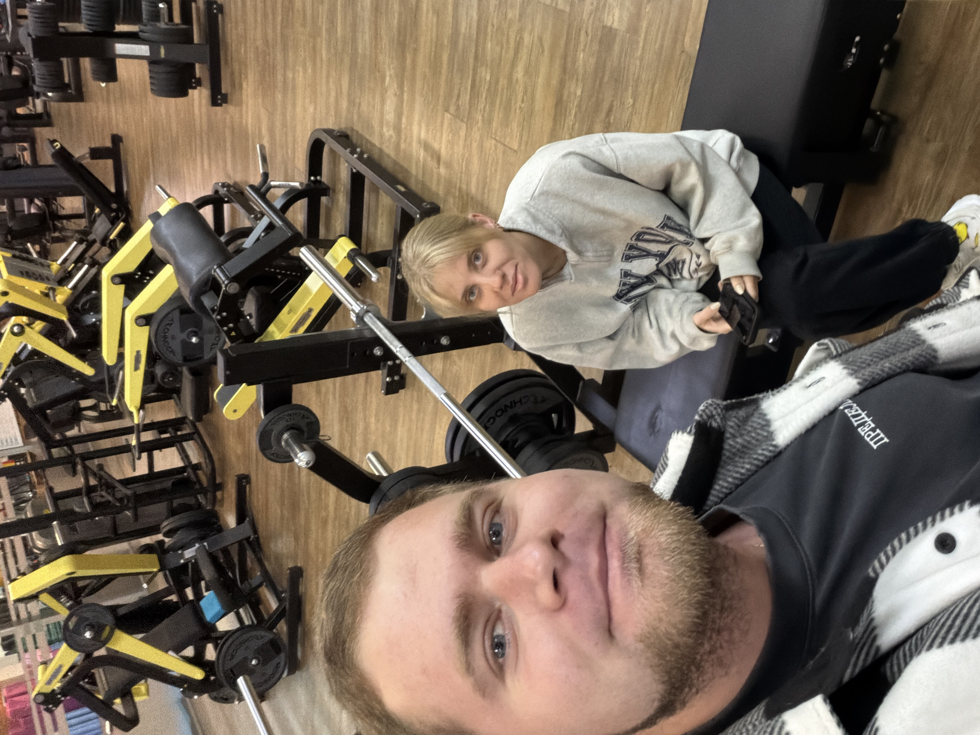 The image appears to be a selfie taken in a gym setting. A man with blue eyes and a beard is in the foreground, wearing a black shirt under a white and black plaid flannel. Behind him and slightly to the left, a woman with blonde hair is sitting on a weight bench, wearing a light gray sweatshirt and black pants. She is holding a black object, possibly a phone or remote. The gym has wooden-look flooring, and in the background, there are various weightlifting machines, some with yellow accents. Based on the presence of "Technogym" branded weights, and the location of Chaikovsky, Russia, it is likely this photo was taken in a local gym there.