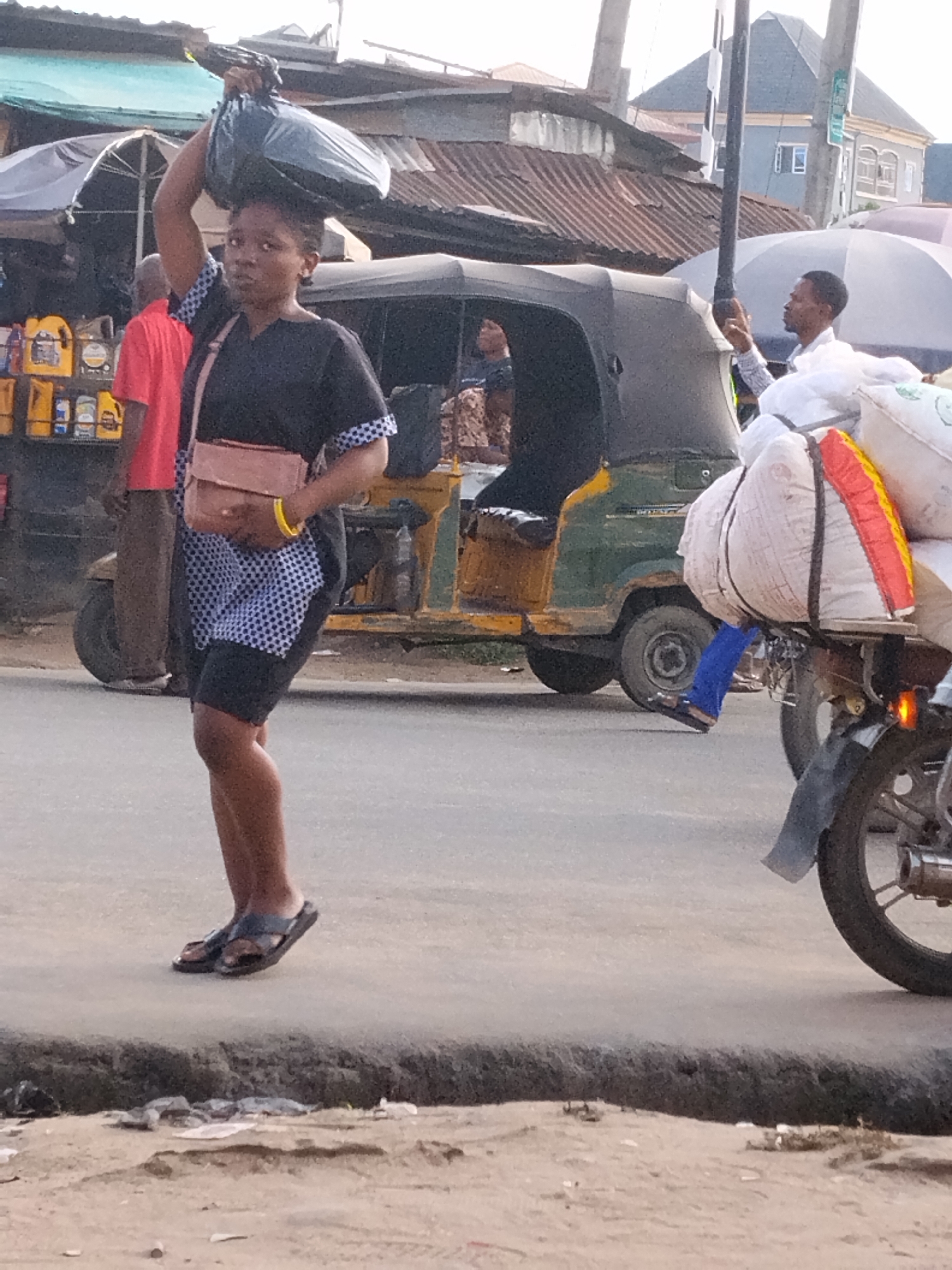 A young woman, dark-skinned, is carrying a large, black bag on her head and walking on a paved road in Aba, Nigeria. She wears a dark-colored, patterned dress with black shorts underneath, brown sandals, a small brown shoulder bag, and a yellow bracelet. Behind her is a "keke," a three-wheeled taxi. To the right, a motorcycle loaded with large white bags is partly visible. Further back, a man with a white umbrella stands next to the motorcycle. A man in a red shirt stands near a display of motor oil in the background. Buildings with corrugated metal roofs and utility poles are also visible. The road edges are unpaved with scattered debris.