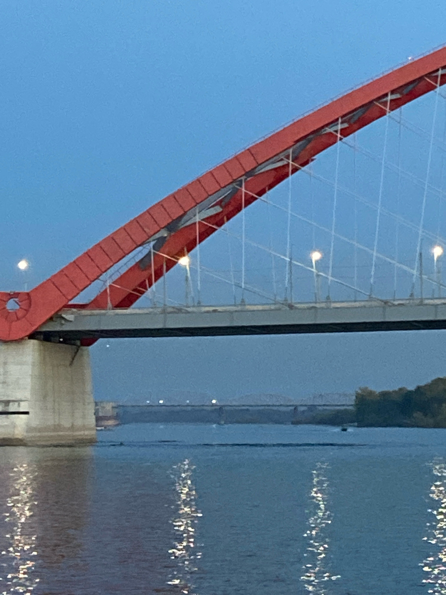 The image shows a view of the Bugrinsky Bridge in Novosibirsk, Russia. The bridge, painted a vibrant red, spans a wide, calm river. The sky is a deep blue, indicating either early morning or late evening twilight. The bridge's structure, with its distinctive arch and supporting cables, is prominently featured in the foreground. The river below reflects the lights of the bridge and the sky, creating a shimmering effect on the water's surface. In the distance, another bridge can be seen, along with a forested shoreline and a small boat on the water. The scene is peaceful and serene, capturing the grandeur of the bridge against the backdrop of the natural landscape.