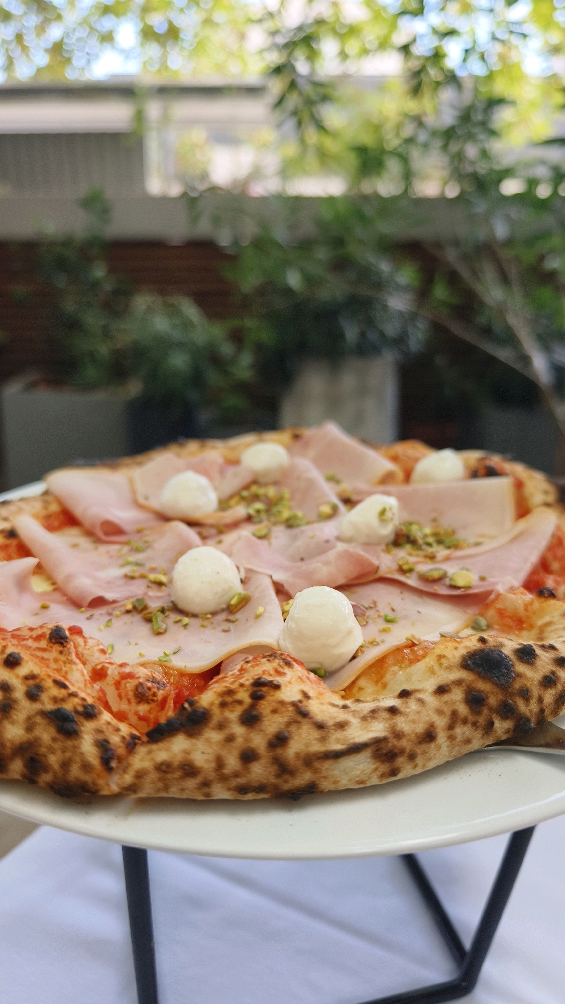 An overhead, close-up shot shows a gourmet pizza, likely Neapolitan-style, with a slightly charred, puffy crust. The pizza is topped with thinly sliced ham, small white balls of fresh mozzarella, and a scattering of chopped green pistachios. The background is softly blurred, suggesting an outdoor setting with greenery and dappled sunlight, possibly a courtyard or terrace in Vienna, Austria, as indicated by the ambient lighting and foliage. The pizza rests on a white plate, which is positioned on a table covered with a white tablecloth.