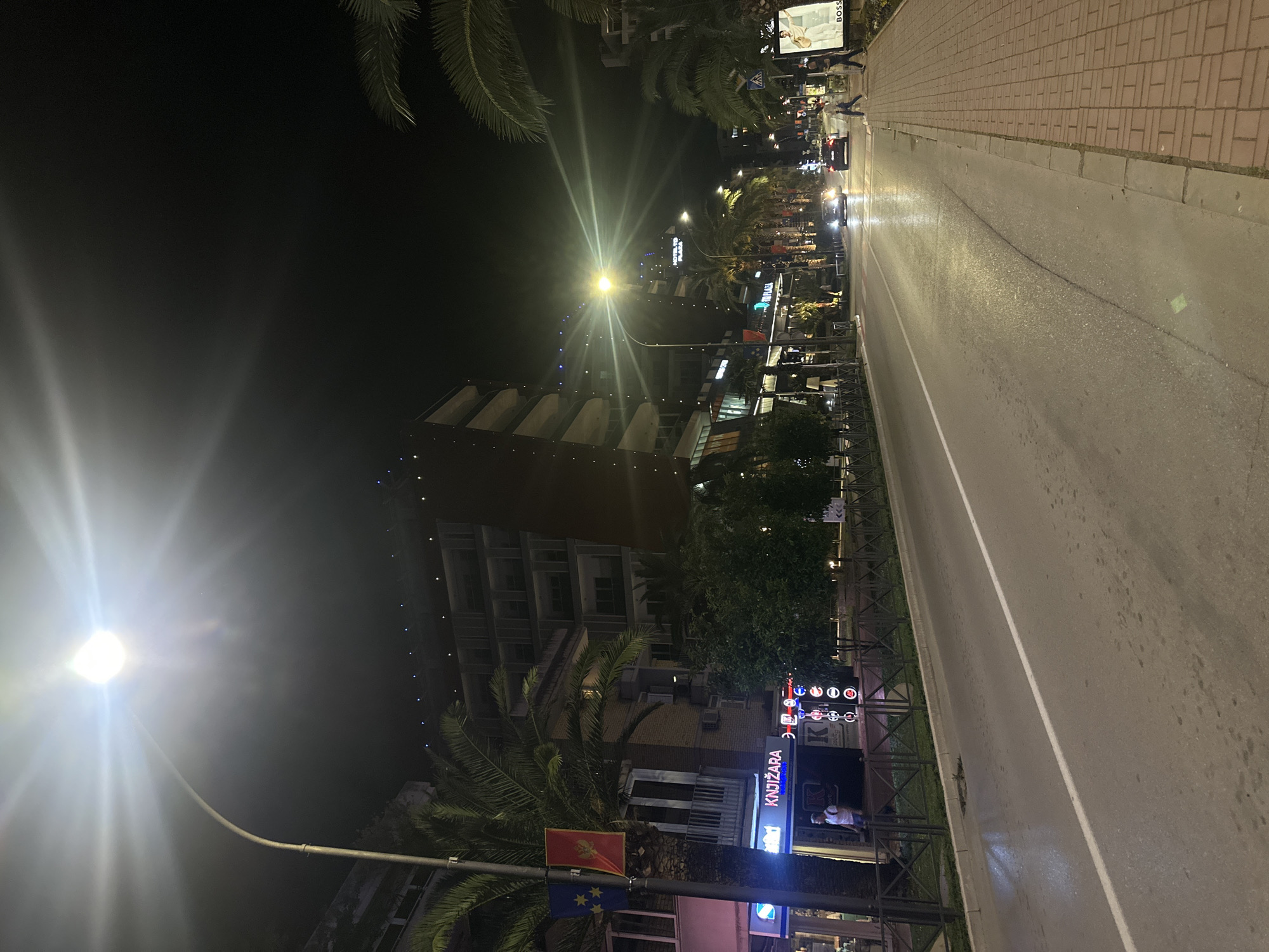 The image depicts a nighttime street scene in Budva, Montenegro. A modern building with a unique geometric design rises in the background, illuminated by string lights. In the foreground, palm trees frame a street lined with shops, including a bookstore named "Knjizara." A streetlamp casts a bright glow, while a car drives down the road. National flags (Montenegro and European Union) are attached to a street light post. Sidewalk is present with few people walking on it.
