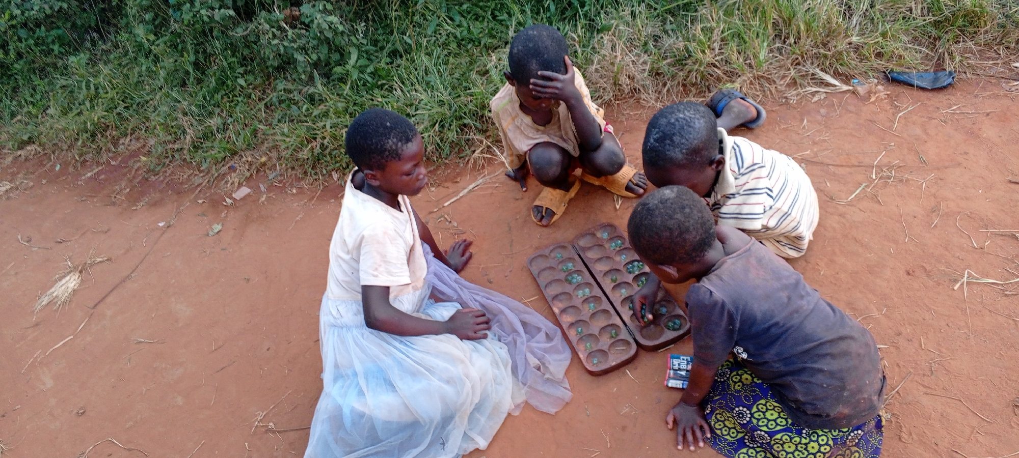 Four African children are playing a traditional board game, likely Mancala, on a red dirt surface in Ngalula, Democratic Republic of the Congo. The board is rectangular with two rows of circular depressions, each containing small, round objects, possibly marbles or stones. The children are positioned around the board, engaged in the game. One child wears a white t-shirt and light blue skirt. Another wears a light-colored shirt and is squatting. A third child is leaning over the board with a dark shirt and patterned pants. The fourth child has their hand on their head and wears sandals. Debris and vegetation are visible in the background. A blue sandal is seen at the right of the frame. A box is visible on the ground between the player wearing the dark shirt and the board.
