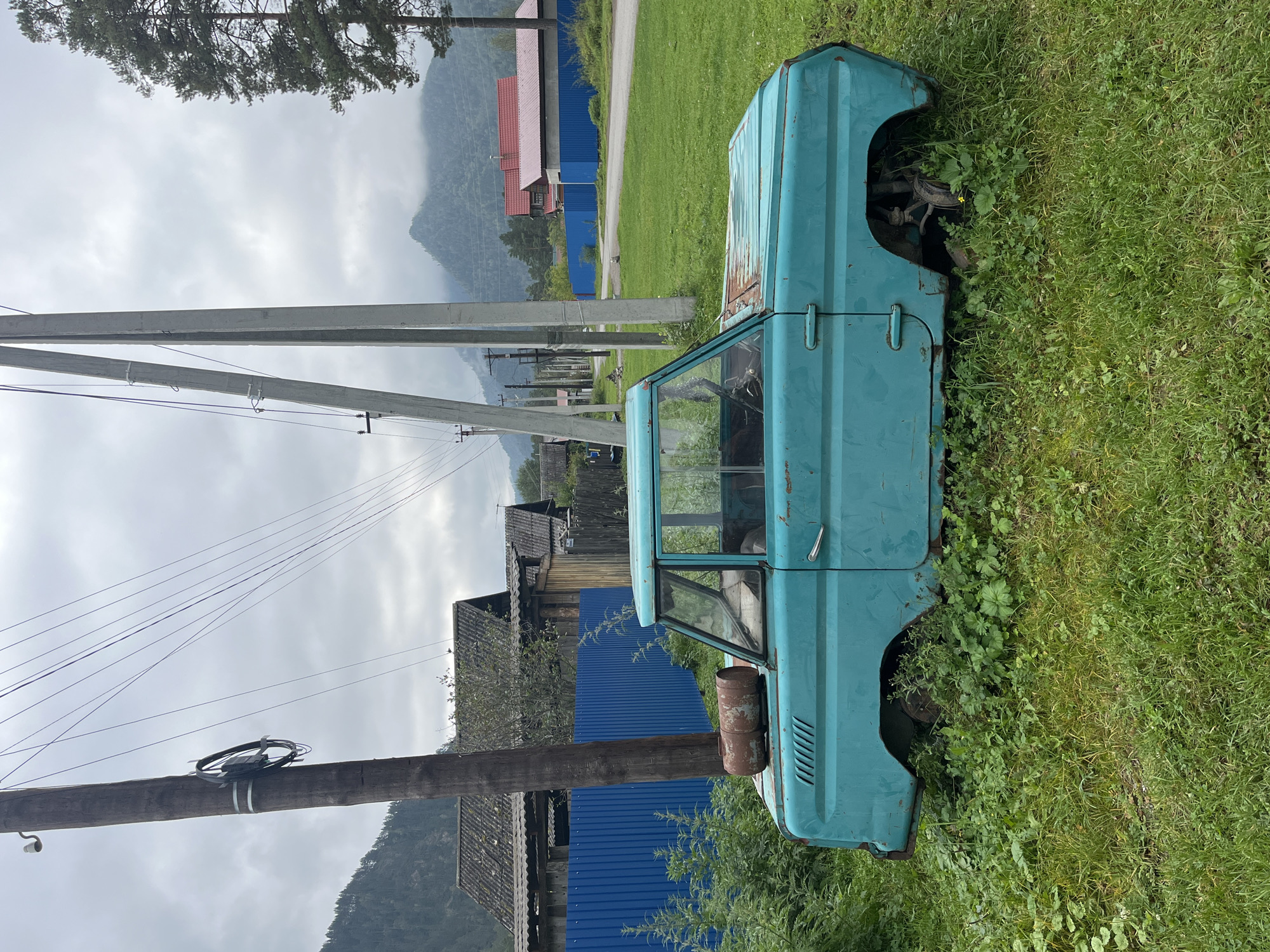 An old, teal-colored car, heavily rusted and overgrown with grass, sits abandoned in a grassy area in Ust-Sema, Russia. The vehicle appears to be a pickup truck with a damaged cab and missing wheels. In the background, traditional wooden houses with dark, weathered roofs line a dirt road. A bright blue metal fence runs along the property. Power lines stretch across the cloudy sky, supported by wooden and concrete poles. Distant, mist-covered mountains frame the rural landscape. The scene is overcast, suggesting a cool, damp day.