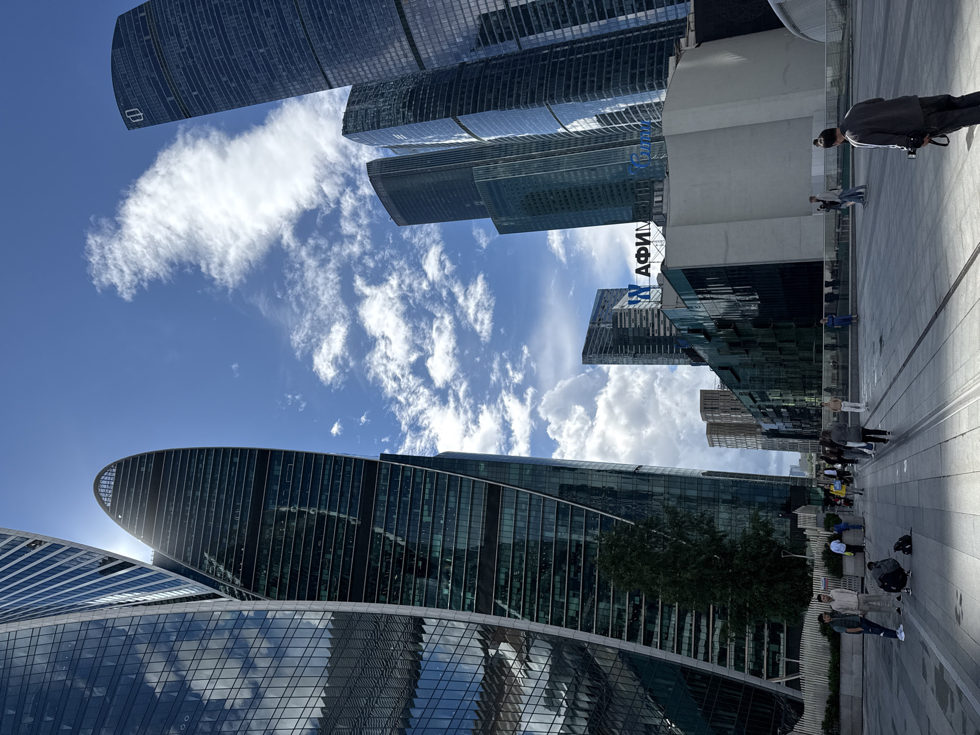The image captures a view of the Moscow International Business Center, also known as Moscow City, a commercial district in western Moscow, Russia. Dominating the scene are several towering skyscrapers with modern, glass facades, reflecting the bright blue sky and scattered white clouds. In the foreground, a paved plaza area is populated by a few individuals. One person is crouched down, seemingly adjusting a bag, while others stand or walk in small groups. A man in the lower right corner is holding a camera, facing away from the viewer. Buildings in the middle ground include a structure with prominent signage reading "АФИМОЛЛ" (AFIMALL), a large shopping mall in the area. The architecture is characterized by sleek lines and impressive height, indicative of a major urban center.