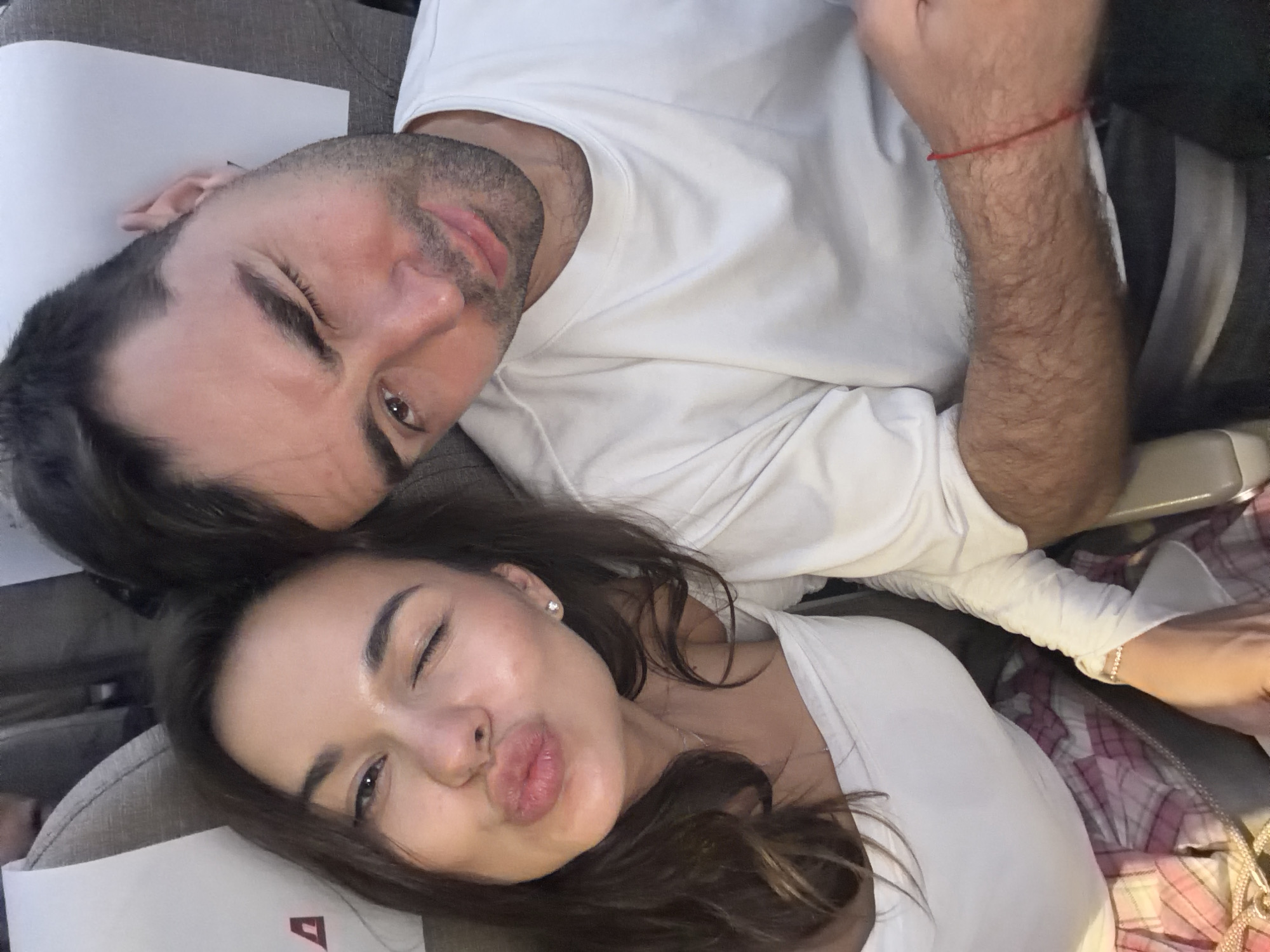 A photograph shows a man and woman lying close together, appearing to be on an airplane or in a confined space with airplane seats and documents present. The man is on the upper left, with short dark hair, wearing a white shirt and a red bracelet on his right wrist. The woman is on the lower right, with long brown hair, wearing a white top, and blowing a kiss at the camera while winking. The environment is dimly lit, and the context suggests travel. The city of Zhangjiajie, China, is not visible or implied in the image.