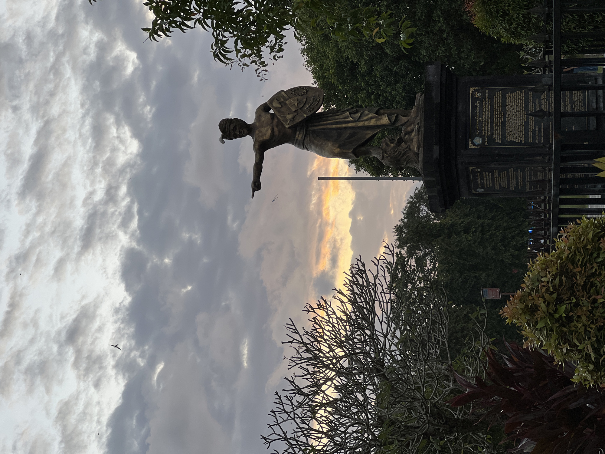 The image shows a bronze statue of a male figure holding a shield and pointing forward, located in Denpasar, Indonesia. The statue is positioned on a dark pedestal with inscriptions. In the background, there are trees and a cloudy sky with hints of sunset colors. Several birds are visible in the sky. The foreground features foliage and a black metal fence. The overall scene suggests an outdoor public space, possibly a park or monument area, at dusk.