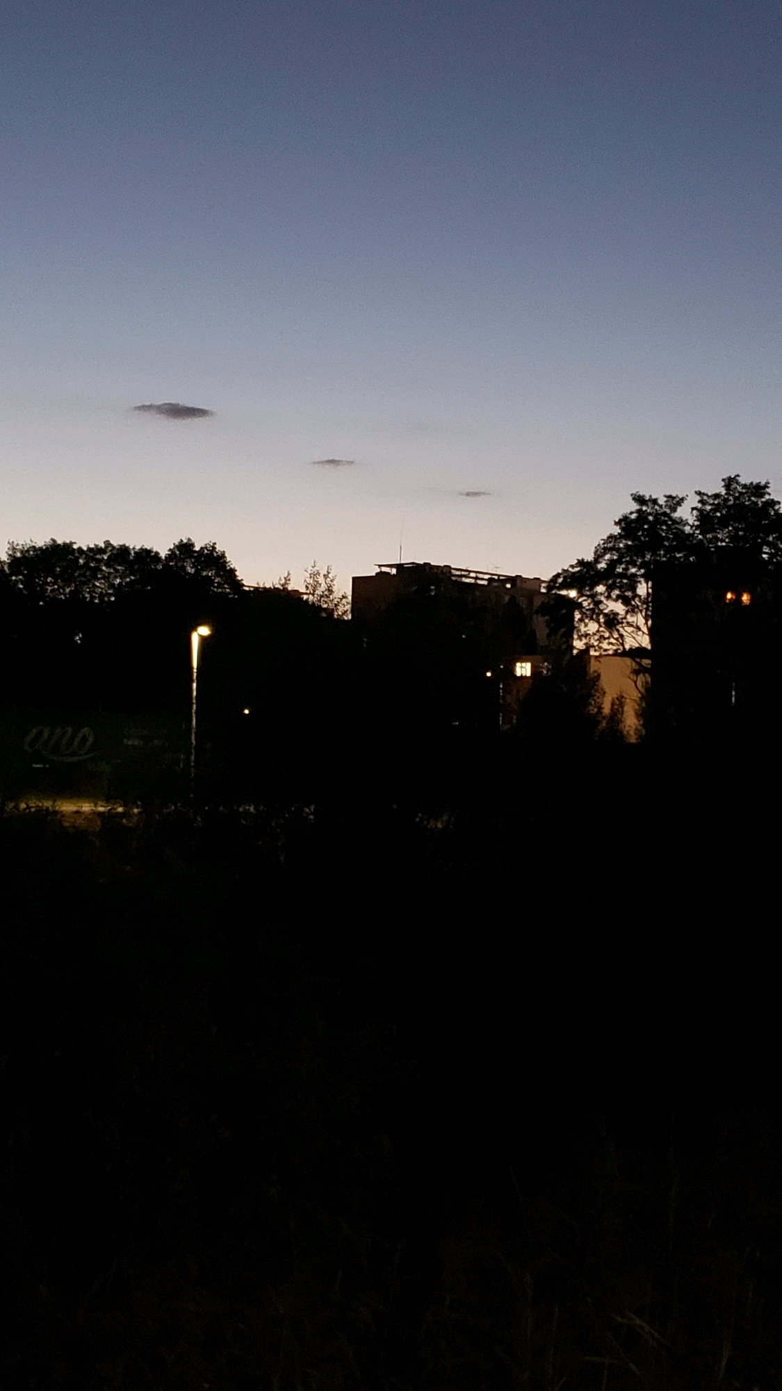 The image depicts a silhouetted landscape at dusk or dawn in Prešov, Slovakia. The sky is a gradient of deep blue fading to a lighter hue near the horizon, with a few wispy clouds. In the foreground, dark foliage and vegetation create a dense, shadowy mass. A single, illuminated street lamp stands prominently on the left side, casting a warm glow. To the right of the street lamp, partially obscured by trees, is a building complex with some windows lit, suggesting occupancy. Faint lettering, possibly a sign, is visible on the left, appearing to read "ano". The overall impression is one of a quiet, urban edge transitioning into night.
