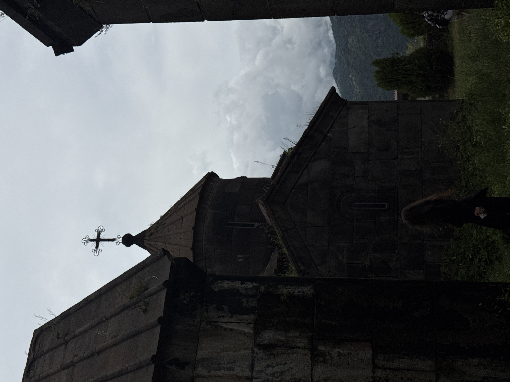 A medium shot captures a view of the Haghpat Monastery in Haghpat, Armenia, under an overcast sky. The stone structures of the monastery are dark and ancient, with one building featuring a conical roof topped by an ornate cross. Vegetation grows on and around the stone walls. In the foreground, a person with long, dark hair, wearing a dark outfit, walks away from the camera, holding a small white object in their right hand. The background shows a lush, green mountainside under a cloudy sky.