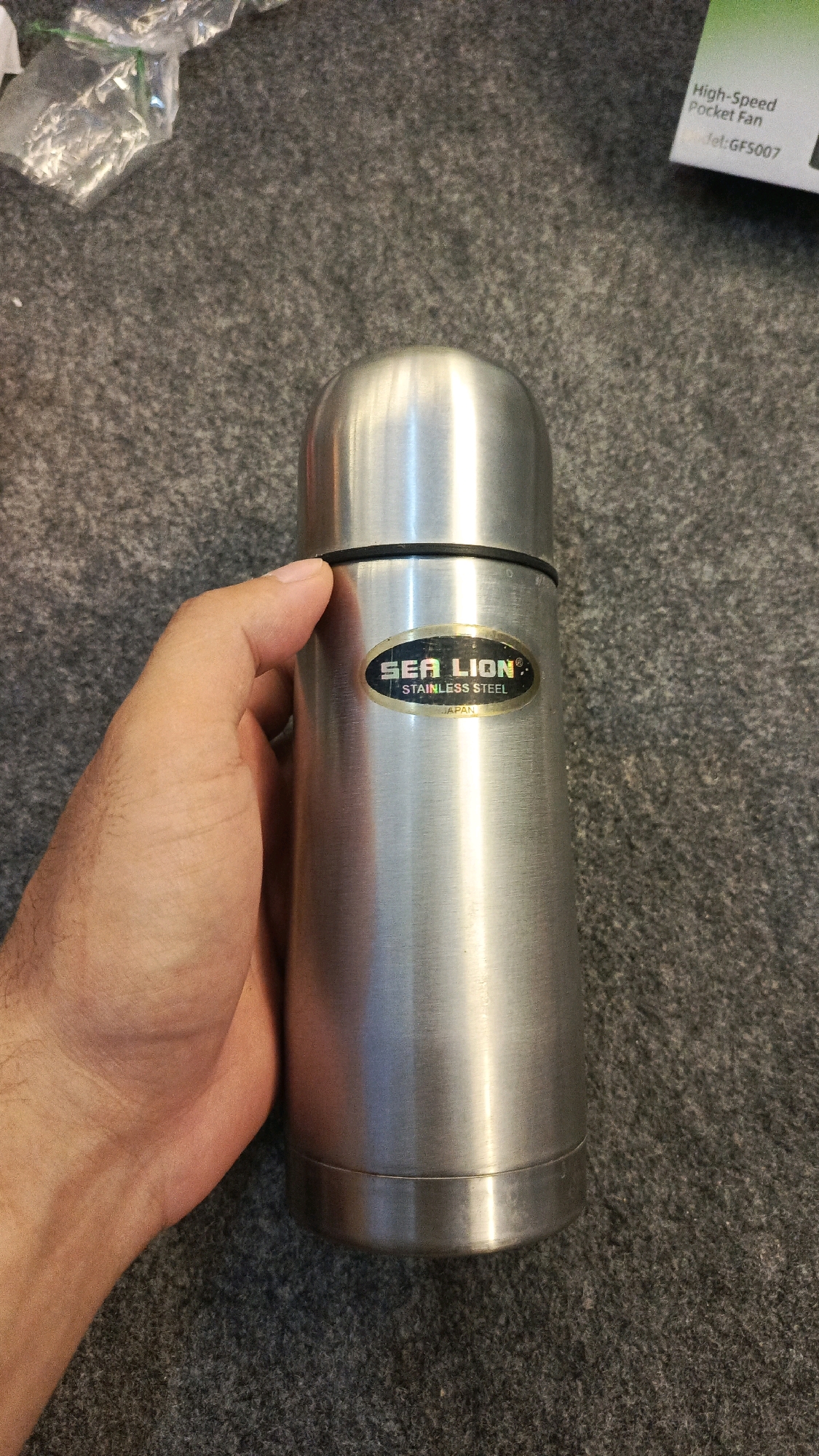 A hand is holding a stainless steel thermos with a "SEA LION STAINLESS STEEL JAPAN" label. The thermos has a black band around the top. The background is a textured gray carpet. There is also a partially visible white and green package in the upper right corner, with text that includes "High-Speed Pocket Fan" and "Model: GFS007". The image was taken in the city of Babadan, Indonesia.