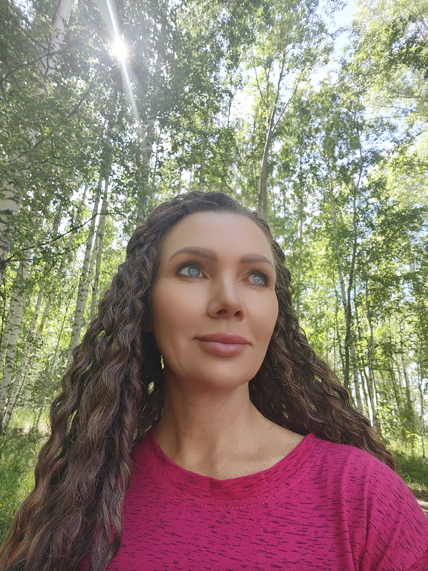 A head and shoulders, outdoor, selfie-style photograph features a woman with long, dark, curly hair and blue eyes. She is wearing a bright pink t-shirt. The background consists of a dense forest with tall, slender trees, possibly birch trees due to their white bark. The sun is shining brightly through the trees, creating lens flare and dappled light. The woman is looking slightly to her right, with a gentle smile on her lips. The overall atmosphere is bright and natural, suggesting a sunny day in a wooded area.