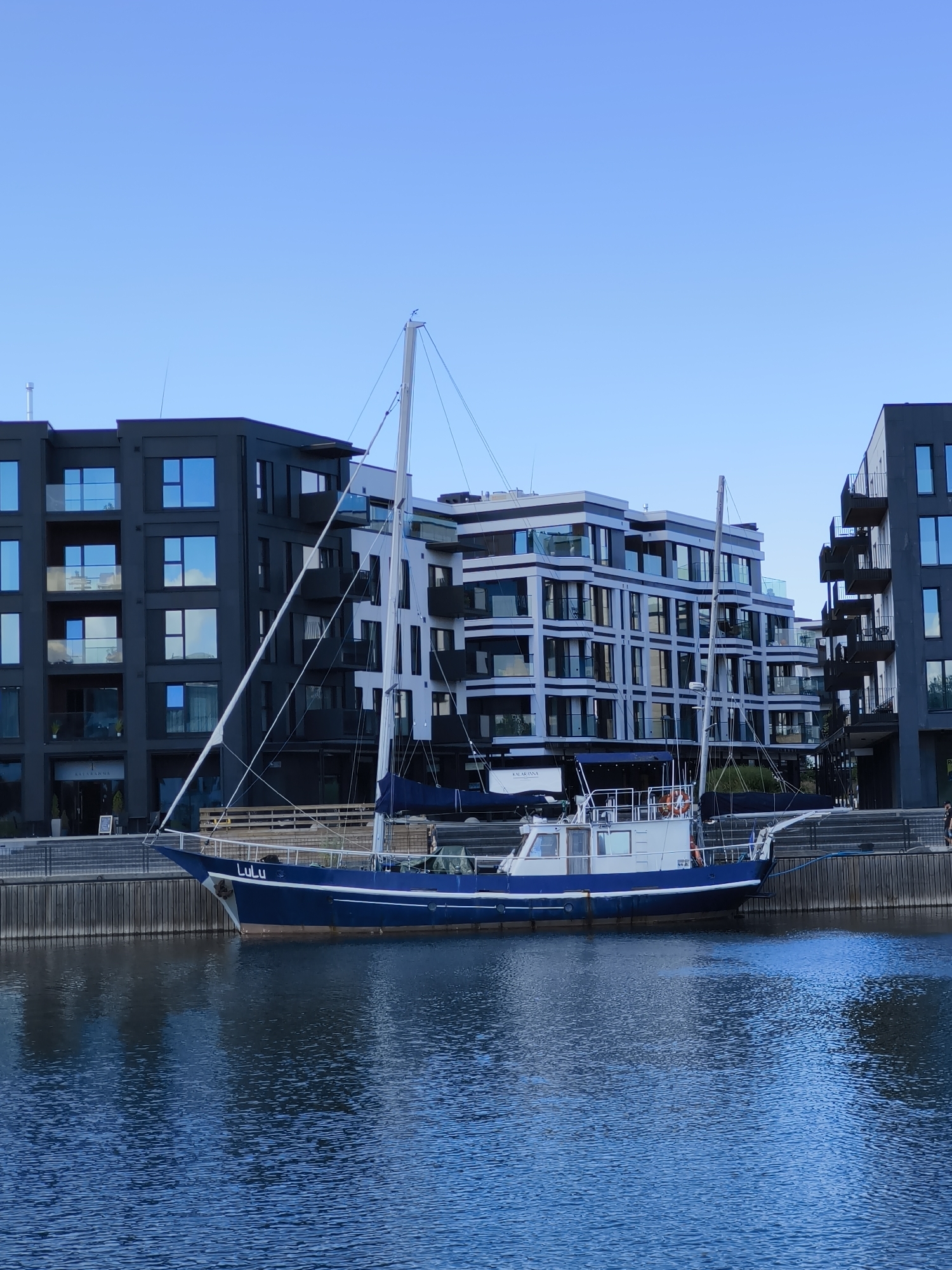 A blue and white sailboat named "Lulu" is docked in front of modern apartment buildings on a clear day. The sailboat is positioned in the foreground, with its hull partially submerged in the blue water. Behind the boat, a wooden pier runs along the waterfront. The apartment buildings are contemporary in design, featuring dark gray and white facades, with large windows and balconies. The sky above is a clear, pale blue, indicating fair weather. The scene appears to be located in a harbor or marina within the city of Tallinn, Estonia, possibly in the vicinity of the Tallinn Old Town or its surrounding developments. There are no visible people interacting with the boat or in the immediate vicinity of the buildings, though a distant figure can be seen walking on the pier.