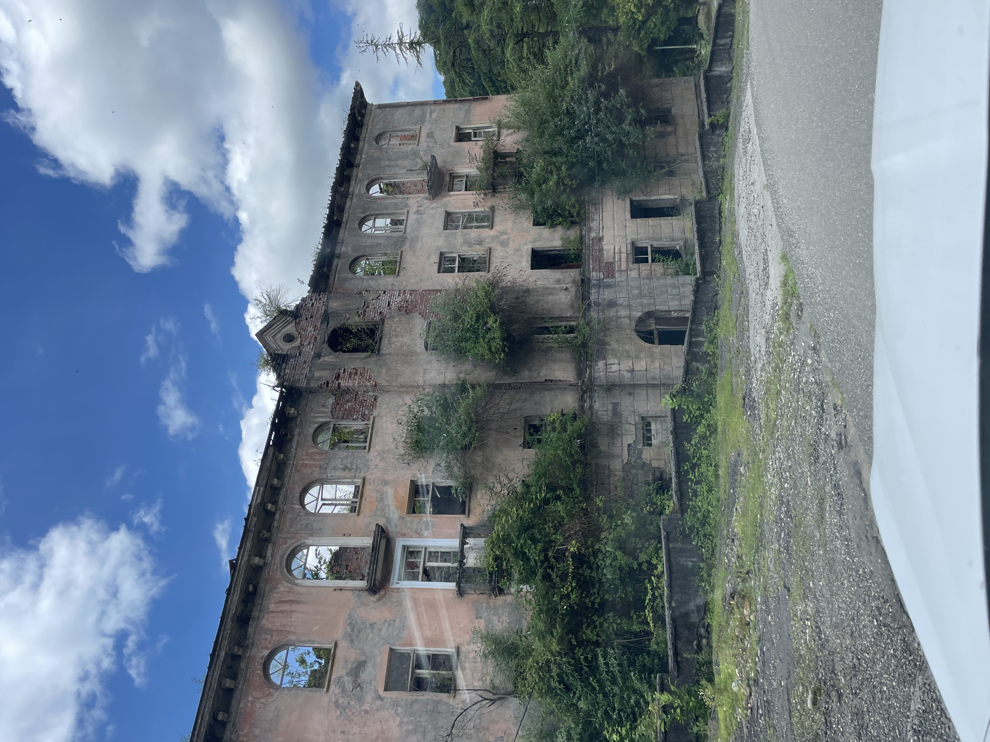 The image shows a large, derelict building in Tqwarchal, Abkhazia. The building is several stories high, with a faded pink facade and numerous broken windows. Vegetation is growing over and around the structure, suggesting it has been abandoned for a significant period. The building appears to be made of stone and stucco, with some exposed brickwork where the facade has crumbled. The architecture suggests it was once a grand edifice, possibly a hotel or sanatorium, with arched windows on the upper floors and balconies. In the foreground, a gravel driveway and part of a white vehicle are visible, indicating the photograph was taken from a car. The background consists of lush green trees and a cloudy blue sky. The overall impression is one of decay and nature reclaiming a man-made structure.