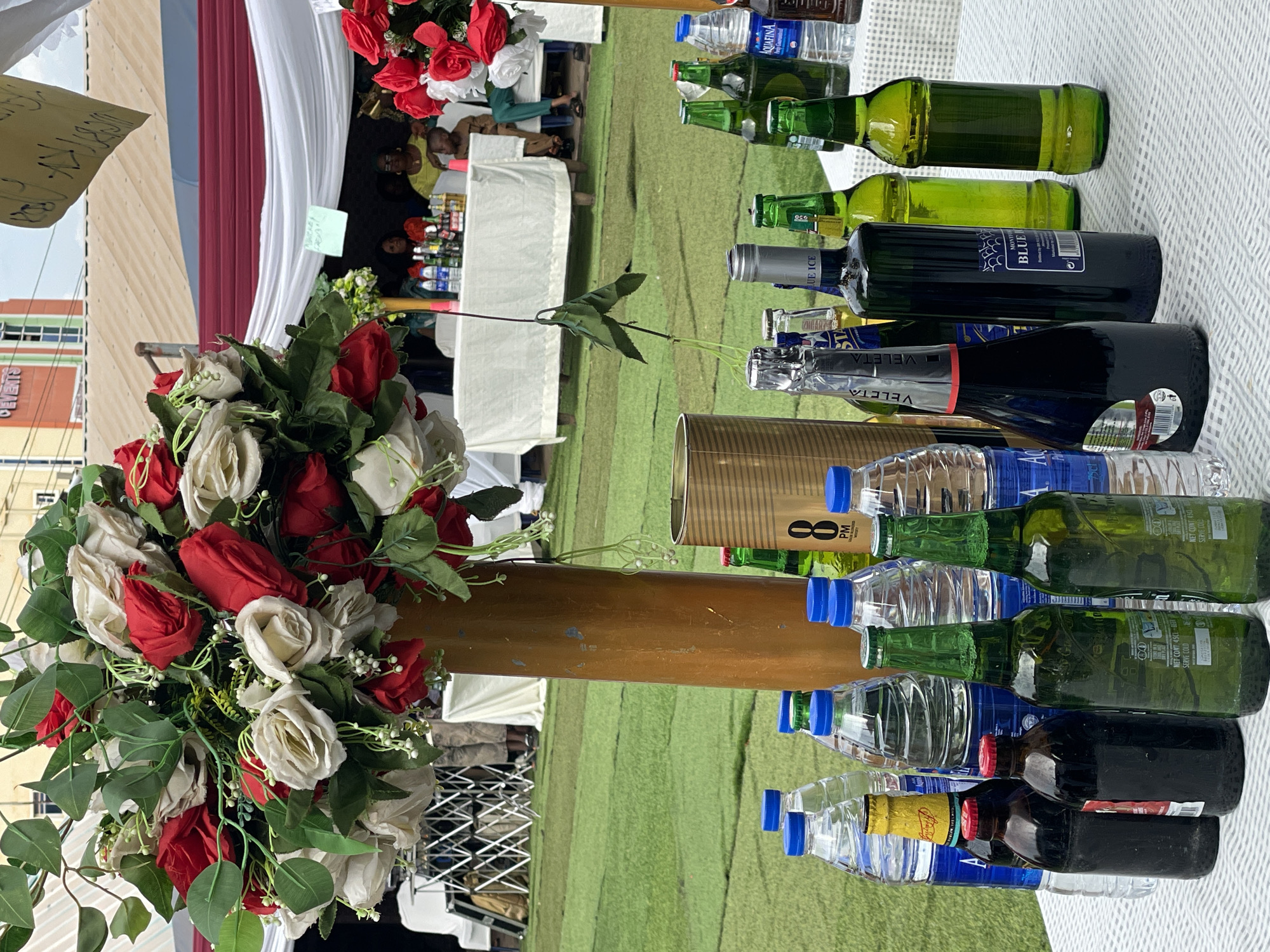 The image depicts a close-up view of an outdoor event, possibly a wedding reception or celebration, taking place in Nkpor, Nigeria. A table covered in a white cloth serves as a focal point, featuring an arrangement of bottles (water, wine, beer) and a tall floral centerpiece with red and white roses in the foreground. In the background, there are visible tables, chairs, and people gathered, indicating a social gathering. The environment appears to be on a lawn. A sign with illegible text is also visible.