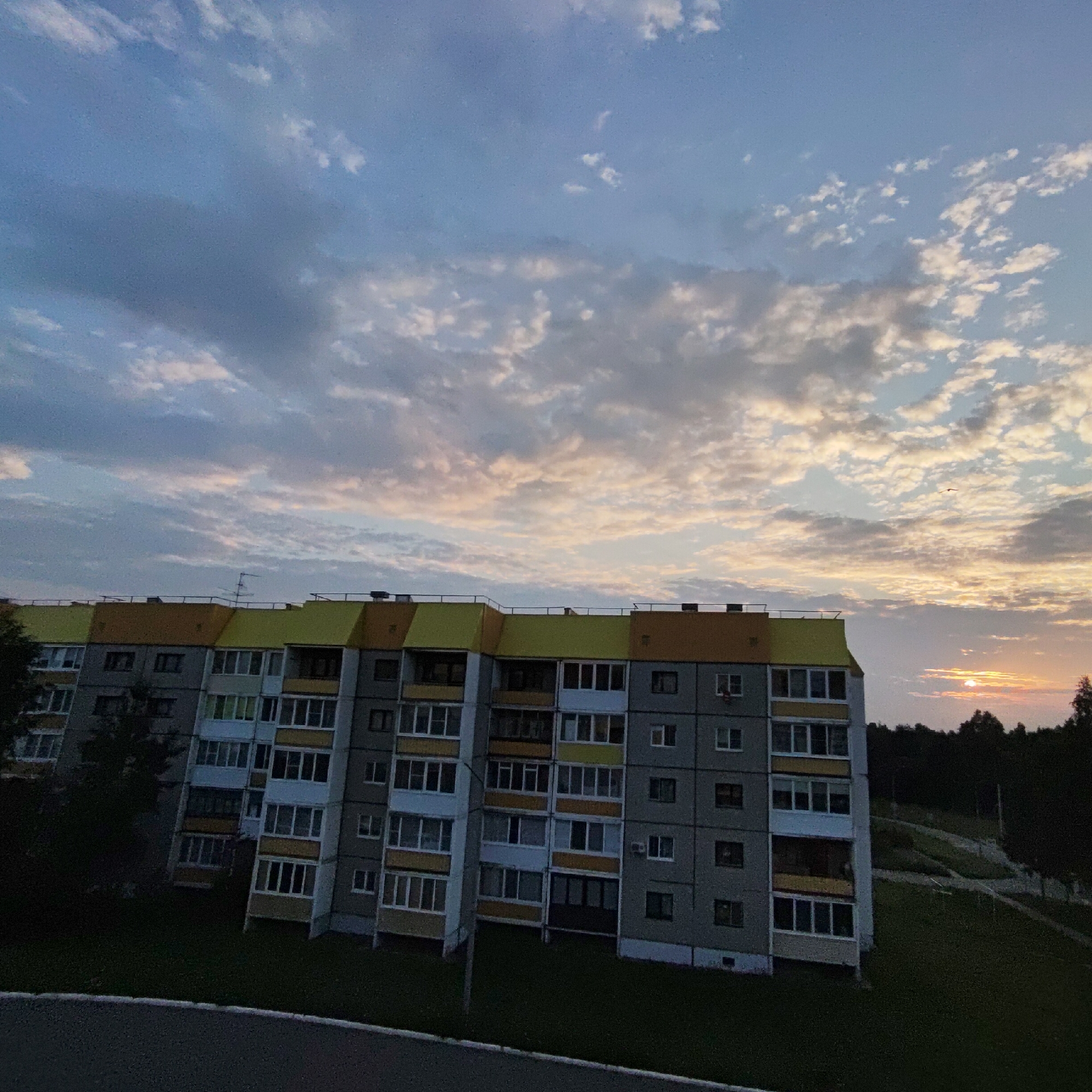 An apartment building in the city of Markovsky, Russia, is pictured against a twilight sky. The building is a multi-story structure with a gray facade and alternating yellow and orange-colored sections at the top. Numerous windows, many with white frames, are visible across the building's front. A satellite dish and railings are present on the roof. In the background, a dense line of dark trees is silhouetted against the sky, and a small, bright sun is visible near the horizon on the right side of the image, casting a soft glow. Scattered clouds, tinged with pink and orange hues, fill the sky. A single bird is visible in flight in the upper right portion of the sky. The foreground shows a dark asphalt surface and a strip of green grass.
