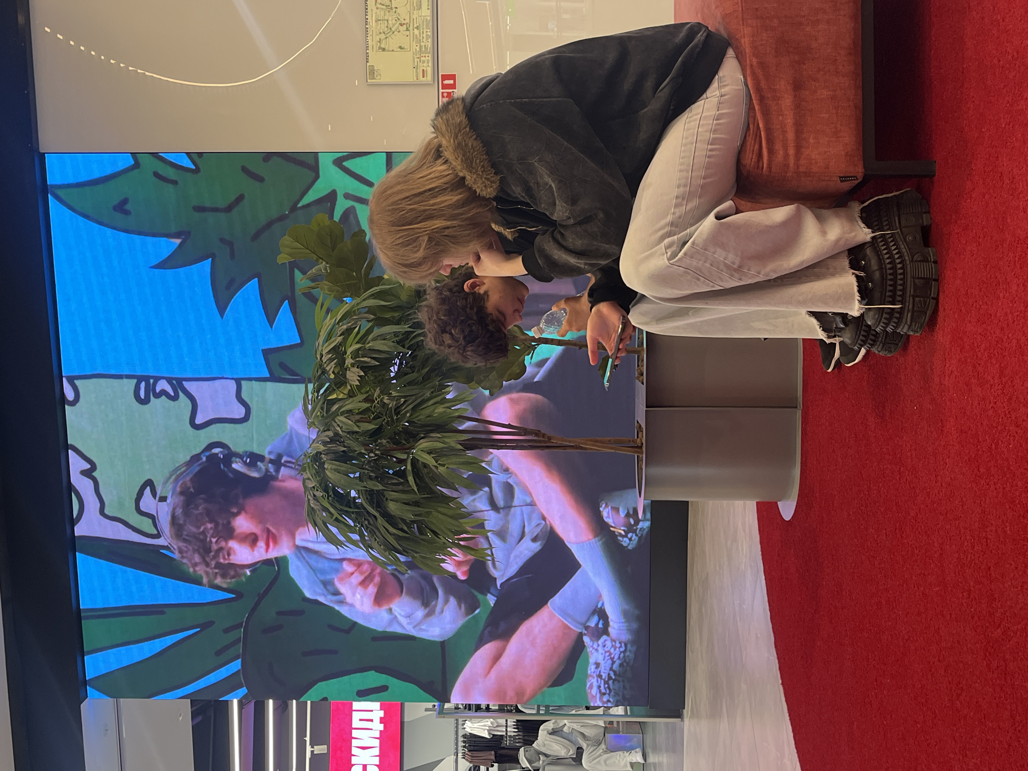 This is an indoor, eye-level shot capturing a scene within a shopping mall in Saint Petersburg, Russia. The background is dominated by a large, vertical LED screen displaying a young man with curly brown hair and over-ear headphones, seated cross-legged in a grey hooded sweatshirt against a stylized blue and green leafy background.
In the mid-ground, a potted plant with green foliage and multiple slender trunks is situated within a grey, multi-sectional planter.
In the foreground, two individuals are seated on a low, rectangular upholstered bench, colored a muted red/orange, which rests on a semi-circular red carpet.
The person on the right, appearing female, has long, light brown hair. They are wearing a dark grey jacket with a brown faux fur collar and light-colored, possibly off-white, loose-fitting pants with frayed hems. Their footwear consists of distinctive, chunky black platform shoes with textured or studded detailing. This individual is leaning forward, holding a smartphone in their right hand and looking down at its screen, with their left hand resting near their chin.
The person seated to their left, appearing male, has dark, curly hair. They are holding a clear plastic bottle to their mouth, appearing to drink from it. Their clothing is mostly obscured by the other individual and the plant.
To the far left of the image, elements of a retail store are visible, including mannequins dressed in light-colored clothing and a bright pink sign with the Russian word "СКИДКИ" (DISCOUNTS). To the far right, a light-colored wall features a white panel with a map labeled in Russian "ПЛАН ЭВАКУАЦИИ ПРИ ПОЖАРЕ" (EVACUATION PLAN IN CASE OF FIRE) and a red fire safety sign. The floor beyond the red carpet is light-colored tile or polished concrete. The scene is illuminated by overhead artificial lighting.