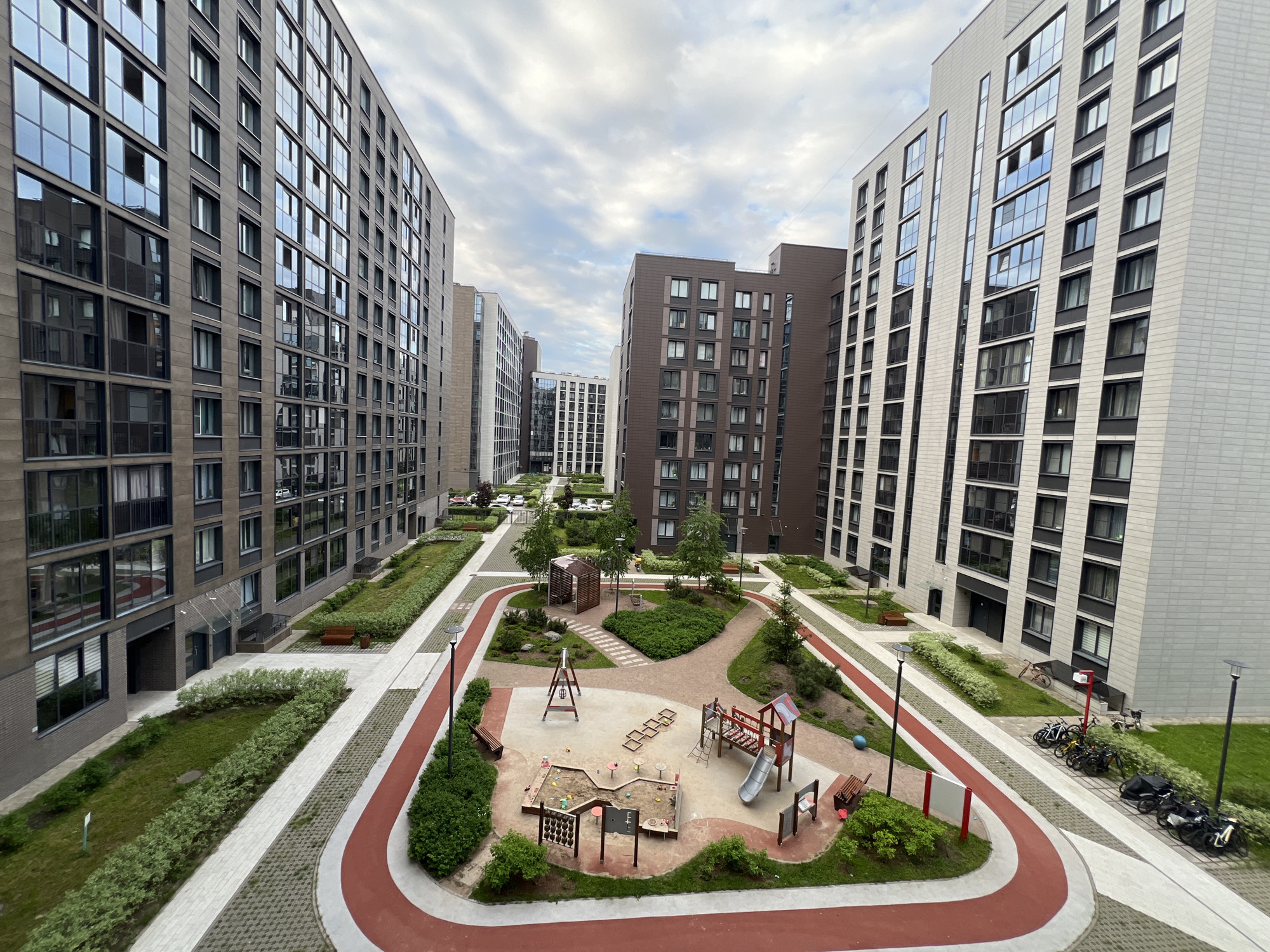 An elevated, wide-angle view captures a modern residential complex courtyard, featuring multiple contemporary apartment buildings arranged around a central green space. The buildings exhibit diverse facades, with some clad in dark gray or brown panels and large, reflective windows, while others feature lighter gray or white exteriors. The architectural style is sleek and functional, common in new urban developments.
The courtyard is meticulously landscaped with manicured lawns, low shrubs, and small trees, defining various zones. Paved pathways of light gray interlocking pavers weave through the green areas, alongside a distinctive recreational track with a red-lined edge, indicating a running or cycling path.
At the heart of the courtyard is a children's playground, equipped with multiple activity structures. This includes a large sandpit area with toys, a slide connected to a playhouse-like structure, swings, and other smaller climbing elements. Several wooden benches are strategically placed within and around the playground for seating. Near the playground, a small wooden pergola or shelter is situated amidst the greenery.
Further along the pathways, street lamps provide illumination. On the right side of the image, near one of the lighter-colored buildings, a cluster of bicycles and a cargo bike are parked. In the background, beyond the immediate courtyard, more residential buildings are visible, suggesting a larger complex, and some parked cars can be faintly seen.
The sky is overcast with scattered clouds, indicating a daytime setting, likely late morning or afternoon. No people are visible, leaving the playground and paths empty. The scene conveys a sense of modern, well-planned urban living with amenities designed for residents, particularly families. No discernible text is visible within the image. The architectural style and communal space design are typical of contemporary developments in cities like Saint Petersburg, Russia.
