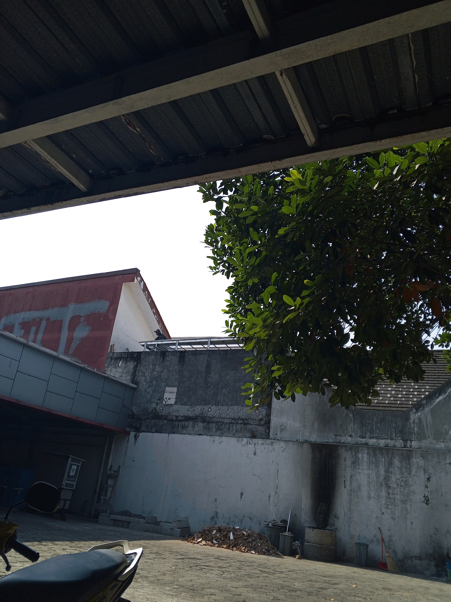 The image depicts an outdoor scene in the city of Condongcatur, Indonesia. In the foreground, the back of a dark-colored motorcycle is visible, with its seat and rear handlebars clearly shown. The ground appears to be paved with greyish interlocking blocks, possibly a parking area or courtyard.
To the left, a building with a grey, angled facade and a red roof with white trim is partially visible. Further back, a tall, light-colored wall runs horizontally across the midground, with some areas showing water stains or dark marks. Above this wall, a section of a building with a grey tiled roof is visible.
On the right side of the image, a large tree with dense green foliage extends from the upper right corner, partially obscuring the sky and parts of the buildings. The sky is bright white, suggesting daytime.
In the background, on top of the light-colored wall, a person wearing a cap and a dark shirt is standing near what appears to be a metal structure. There are also some piles of debris or construction materials on the ground near the wall, along with trash cans and a broom leaning against the wall. The overall impression is an industrial or commercial area with some outdoor space.
