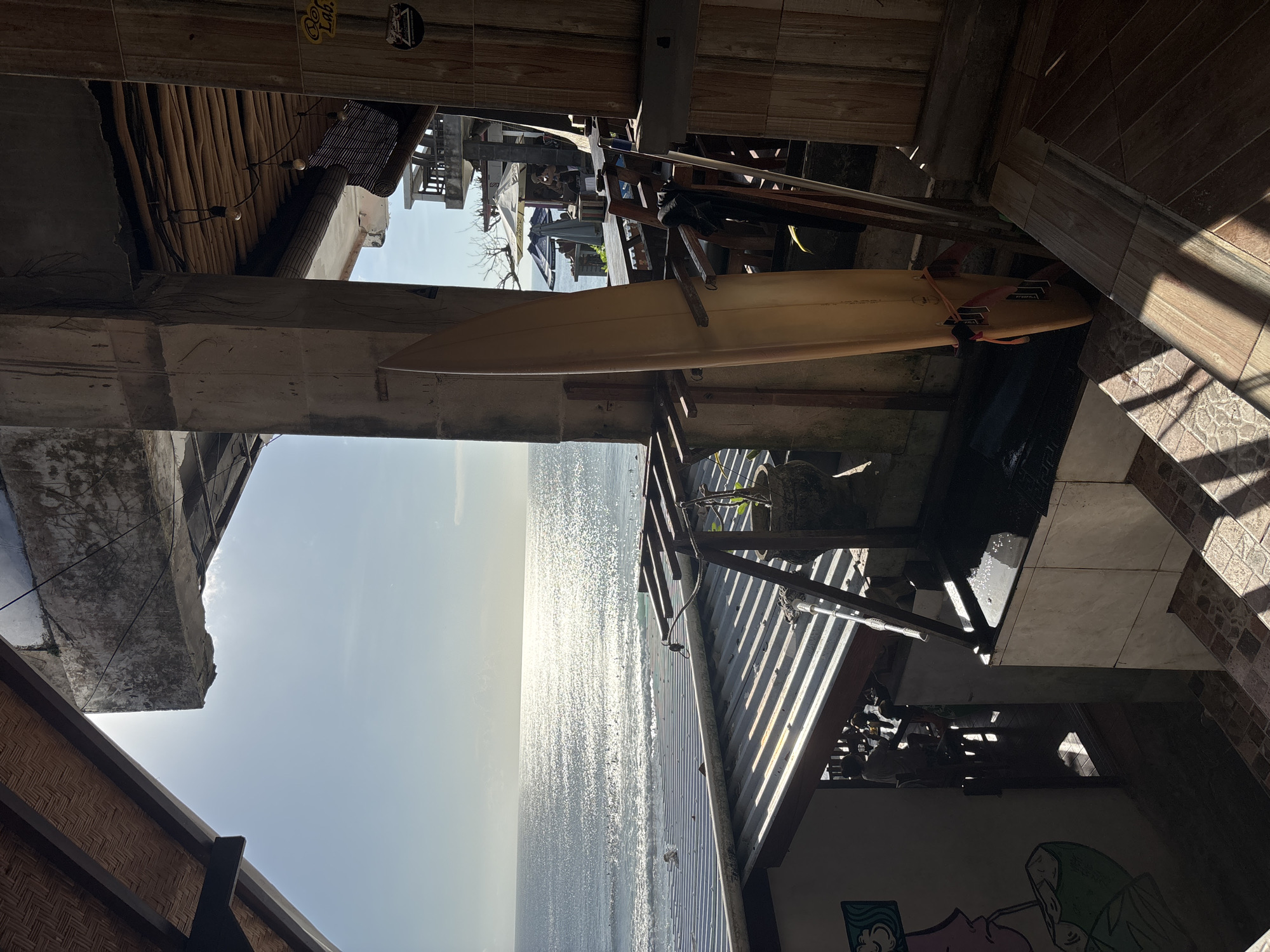 The image shows a partial view of the ocean from what appears to be an elevated or multi-level structure, possibly in Kuta Selatan, Indonesia. A surfboard is mounted horizontally on a makeshift wooden rack or support structure within the building. The foreground includes parts of the building's construction: wooden beams, tiles, and some graffiti-style artwork on a wall. The view through the structure reveals the ocean under a bright, slightly hazy sky. The ocean's surface is reflective, possibly indicating sunlight.