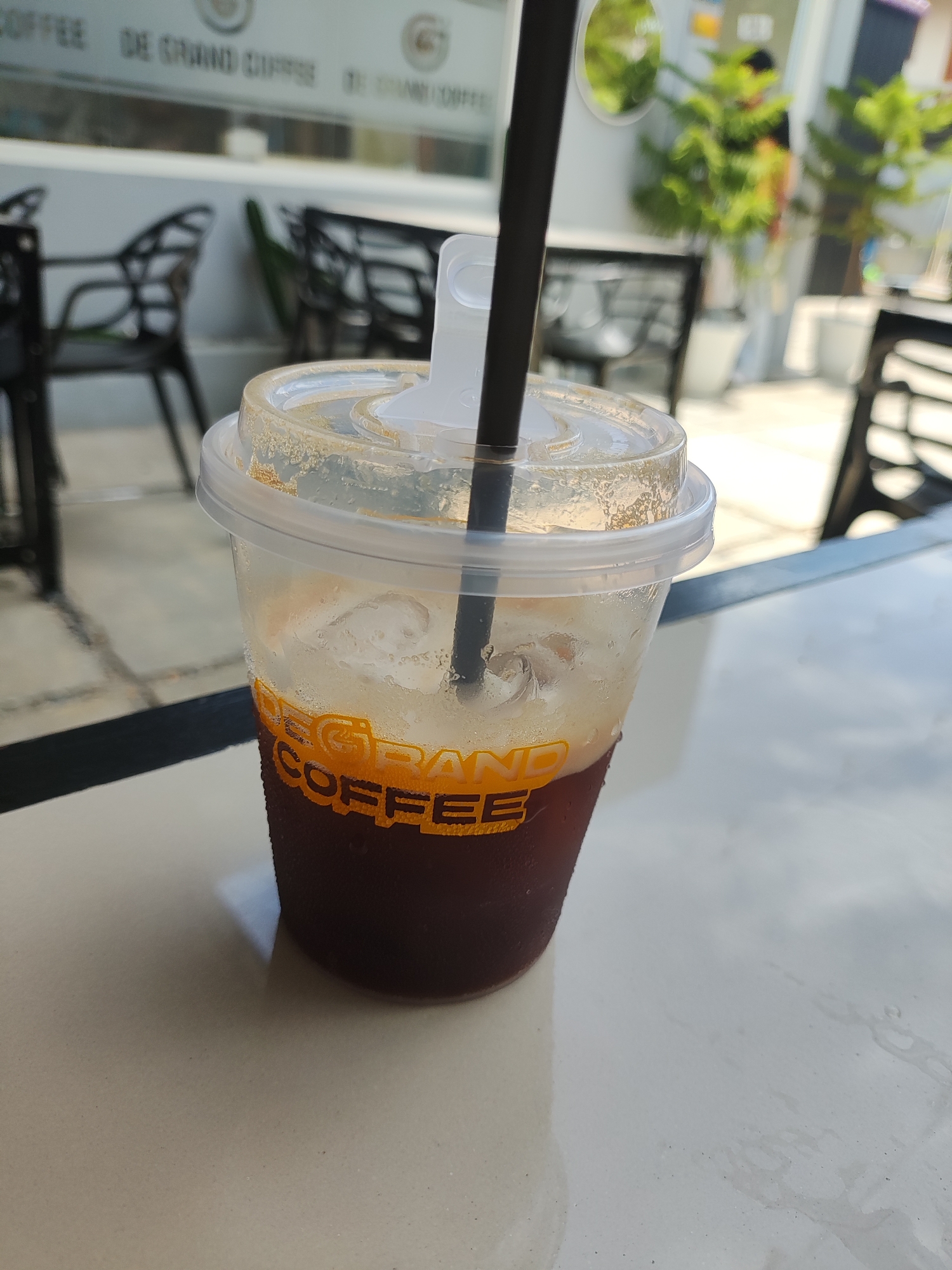 A close-up shot depicts a plastic cup of iced coffee with a black straw, bearing the logo "DE GRAND COFFEE" in yellow text. The cup sits on a white, reflective surface, likely a table. The background shows an outdoor seating area with black chairs, a plant, and a window with the "DE GRAND COFFEE" name displayed. The location is Lhokseumawe, Indonesia, based on the cafe's name on the cup and window. The lighting is bright, suggesting a daytime shot.