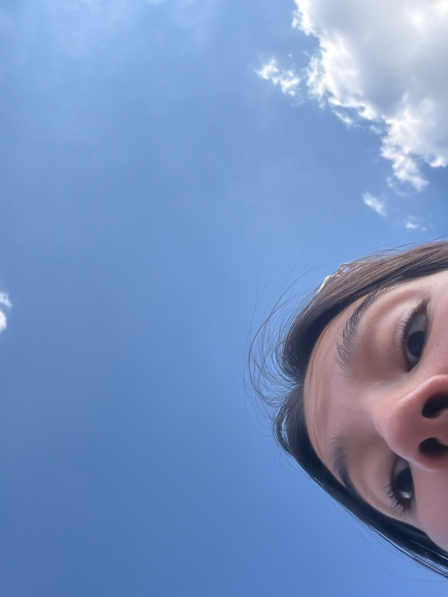 The image is a low-angle, close-up shot of a young person's face looking up at a bright blue sky with scattered white clouds. The subject's right eye, eyebrow, and the bridge of their nose are prominently featured. Their dark hair is partially visible, with some strands blowing in the wind. The lighting suggests it is daytime. There is no identifiable location in the city of San Marcos, United States of America visible in the image; the scene is solely focused on the sky and the person's face.