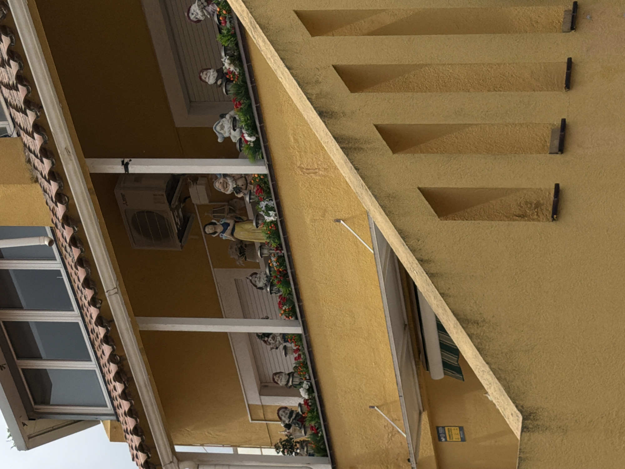 The image shows a building in Sant Andreu de Llavaneres, Spain. The structure has a beige-yellow exterior with a tiled roof and white window frames. The building's architectural details include recessed rectangular shapes in the facade. Along a balcony or ledge, figurines of people and gnomes are arranged amongst greenery and flowers. An air conditioning unit is mounted on an external wall. A sign is visible on a lower part of the building.