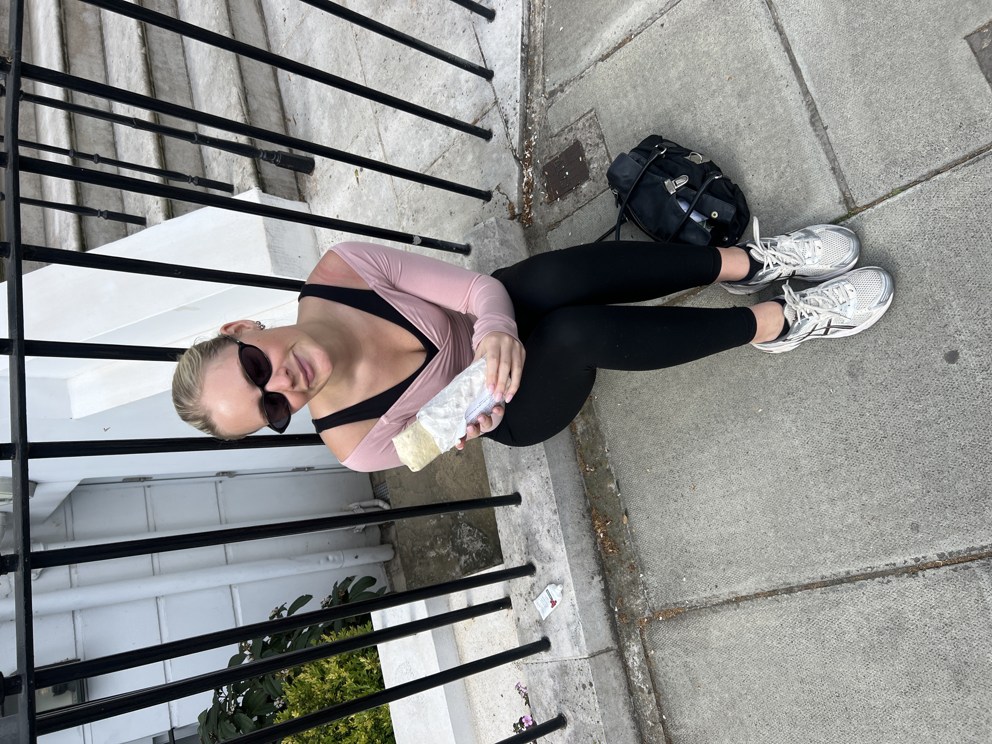 A woman wearing sunglasses, a light pink long-sleeved wrap shirt, and black leggings is sitting on the sidewalk near a wrought-iron fence, holding a wrapped food item, possibly a burrito. Next to her are a black bag and white running shoes. The location appears to be a residential area, possibly in London, United Kingdom, given the architectural style of the buildings and fences.