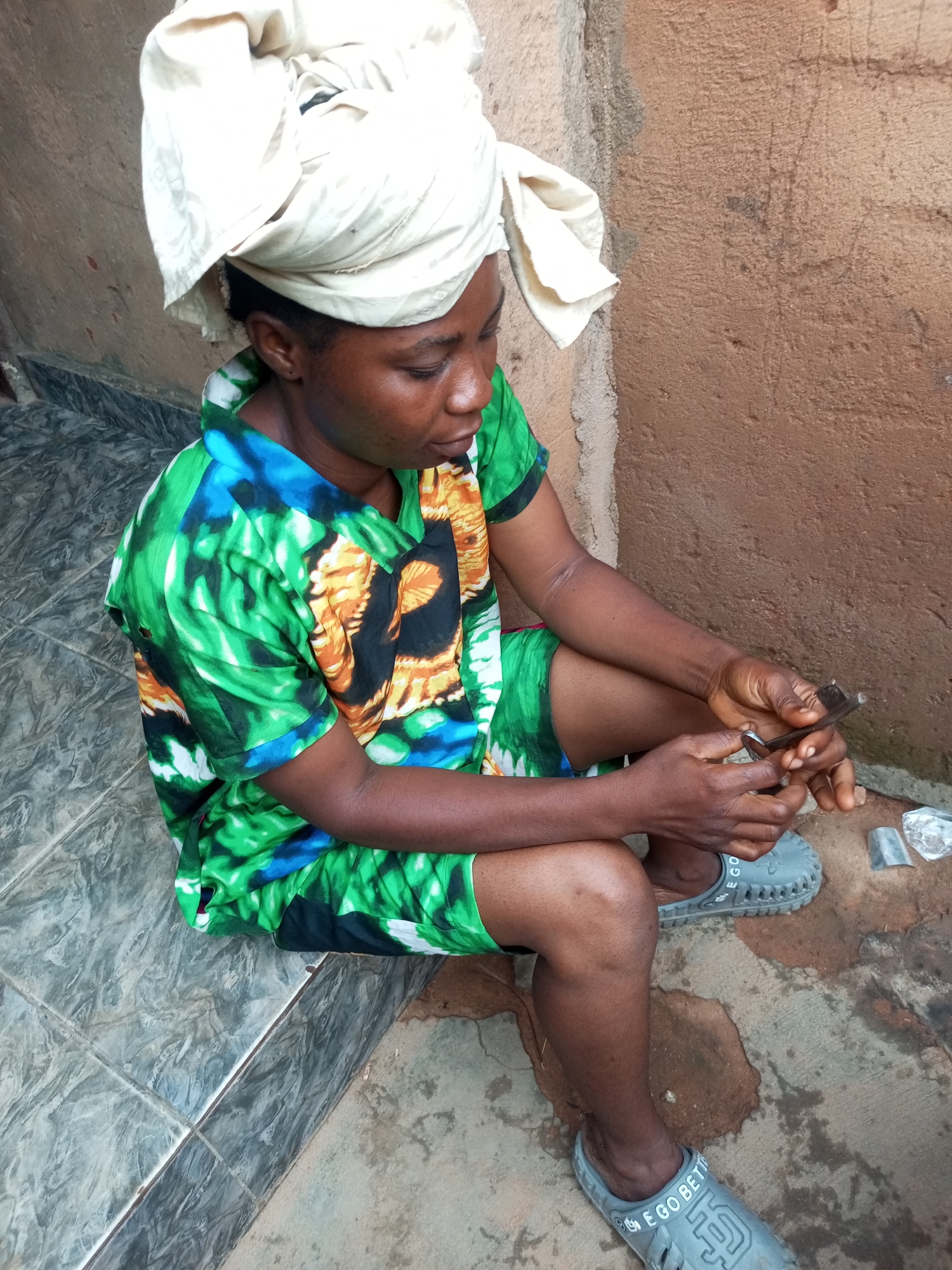 A dark-skinned woman with a white headscarf and a green and blue patterned shirt is sitting on a tiled surface in front of a brown wall. She is holding a small object in her hands and looking down at it. She is wearing grey clogs. The scene is outdoors, and the ground is a mix of tiles and dirt. The wall is rough and textured. There are some small objects on the ground next to her. The image is taken from a slightly high angle.