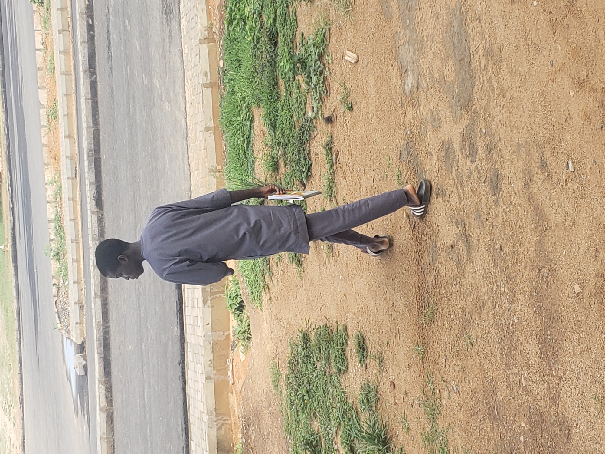 A young person, possibly a male, walks away from the camera on a dirt path near a paved road in Gumel, Nigeria. They are wearing a dark grey traditional outfit, including a long tunic and matching pants, and black and white striped sandals. The person is holding a magazine or booklet in their right hand. The environment consists of a dirt path with sparse green vegetation, leading towards a paved road with a concrete curb and sidewalk. The road appears to be a public street, with some debris and puddles visible. The overall scene is outdoors, during daylight, with overcast or diffused lighting.
