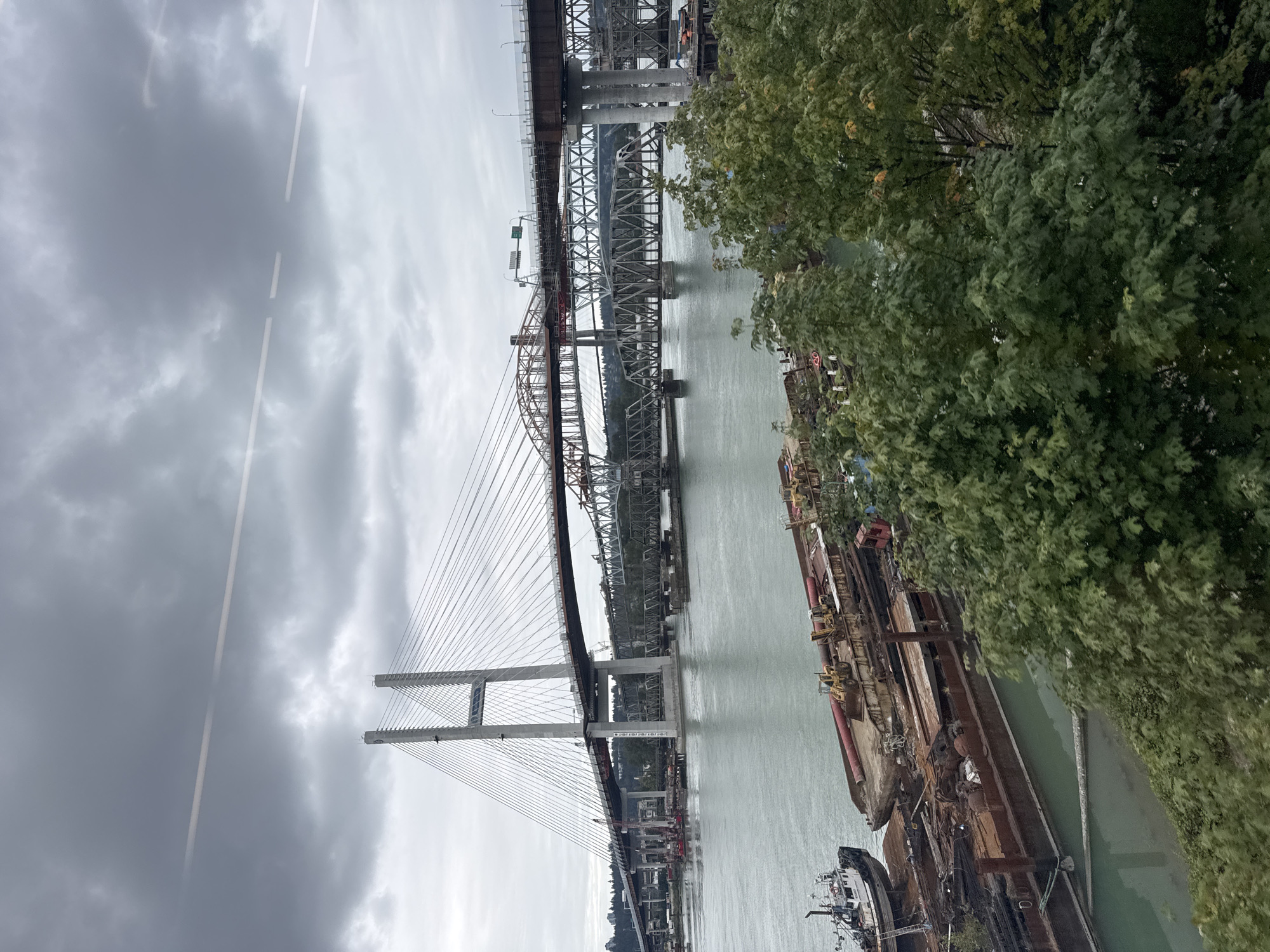 The image depicts a wide river with several bridges spanning across it, with a cloudy, overcast sky overhead. In the foreground, there are several barges docked, some appearing to be used for construction or industrial purposes, with visible heavy machinery like excavators and construction materials. A small tugboat is also present.
The bridges are a prominent feature. The largest and most distant bridge is a modern cable-stayed bridge, characterized by its tall, imposing towers and numerous cables supporting the roadway. Closer to the foreground, there is a steel truss bridge, with a more intricate, lattice-like structure. A third bridge, which appears to be a highway bridge, runs parallel to the truss bridge, with a portion of it being constructed or undergoing renovation, indicated by scaffolding and construction equipment.
The river itself is a muted green color, reflecting the overcast sky. Trees and vegetation line the riverbanks in some areas, particularly in the foreground and background, adding a touch of nature to the industrial and infrastructural landscape. The overall impression is one of urban development and ongoing construction, with a focus on transportation infrastructure over a significant body of water.
This scene is characteristic of the Fraser River in New Westminster, Canada, known for its industrial activity, numerous bridges, and bustling waterways.