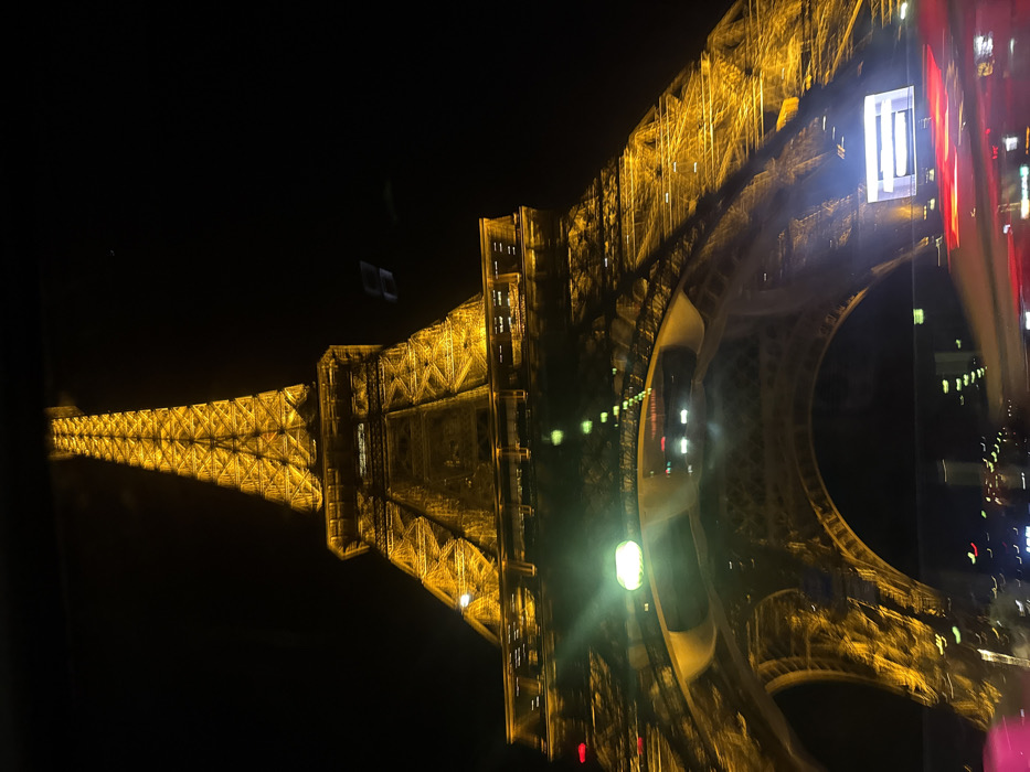 The image presents a low-angle, upward view of the Eiffel Tower in Paris, France, during nighttime. The iconic lattice structure of the tower is prominently illuminated with warm, golden-yellow light, contrasting sharply against a dark, featureless sky. The bottom half of the image exhibits significant light reflections and motion blur, suggesting the photograph was captured from within a moving vehicle. A distinct, elongated green light source is visible centrally in the lower frame, flanked by a linear series of smaller, bright reflections receding towards the left. Horizontal streaks of blurred red and white light occupy the very bottom edge of the image, further indicating vehicular motion. No individuals are discernible. The scene is defined by the stark illumination of the primary subject against the nocturnal backdrop and the dynamic foreground elements.
