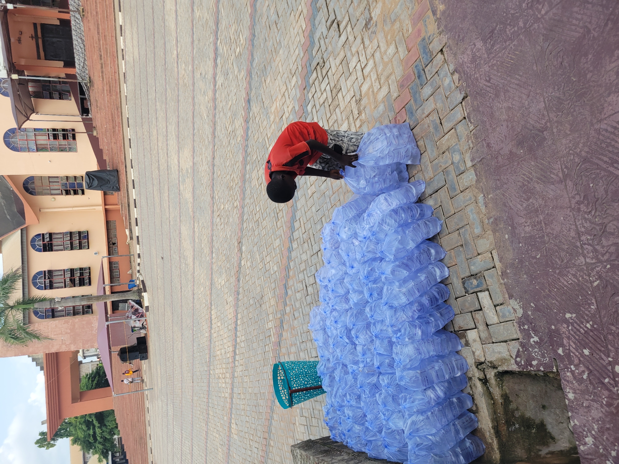 The image depicts a scene in front of a building that appears to be a church or an institution, located in Onitsha, Nigeria. In the foreground, a large pile of blue plastic bags filled with water bottles is stacked on a paved courtyard. A young boy, wearing a red t-shirt and patterned shorts, is attending to the water bottles, picking up a bundle of them. To the left of the water bottles, a blue plastic basket is placed on the ground. The background features the entrance of the building, with steps leading up to it. The building has a light-colored facade and arched windows. Several palm trees are visible in the background, suggesting a tropical environment. The paving stones of the courtyard are a mix of red, grey, and black.