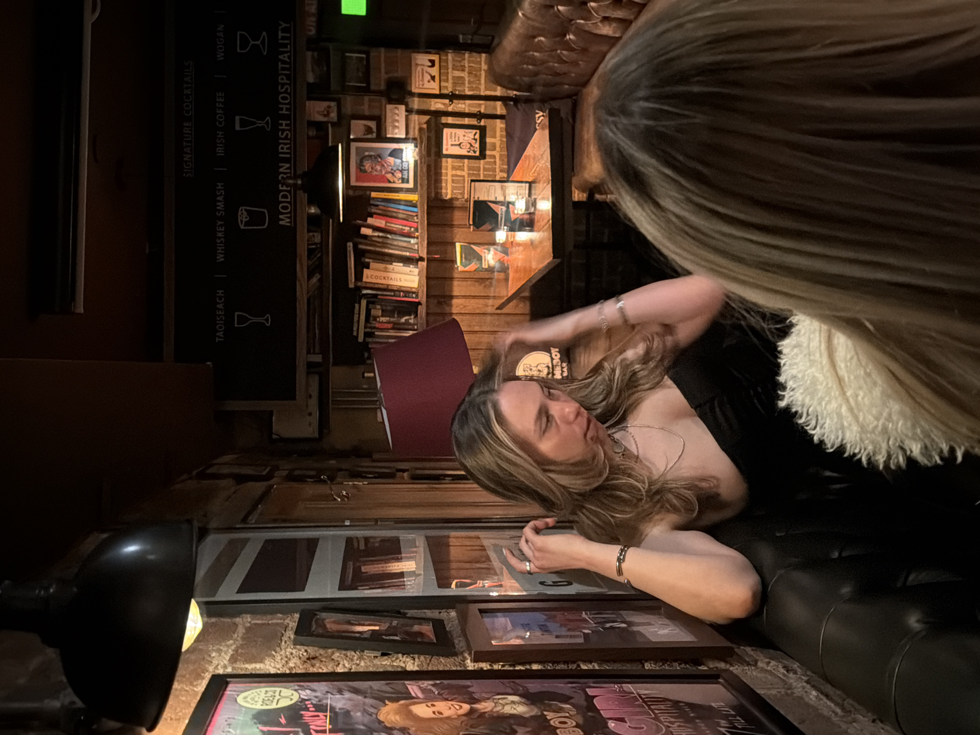 Here is a description of the image:
The image shows the interior of a bar, likely in the city of London, United Kingdom, given the reference to "Modern Irish Hospitality." The space has a rustic feel, with a brick wall decorated with framed pictures and posters. A bookshelf is filled with books, and a black menu board lists signature cocktails like "Taoiseach," "Whiskey Smash," "Irish Coffee," and "Wogan."
A woman with blonde hair is reclining on a black leather couch. Another person, presumably a woman, is partially visible in the foreground with long, blonde hair. The atmosphere is dim and intimate, illuminated by warm lighting from a lamp and other sources. The scene suggests a social gathering in a pub-like setting. The presence of cocktail menus and the bar setting imply this is a place where people are relaxing and enjoying drinks.