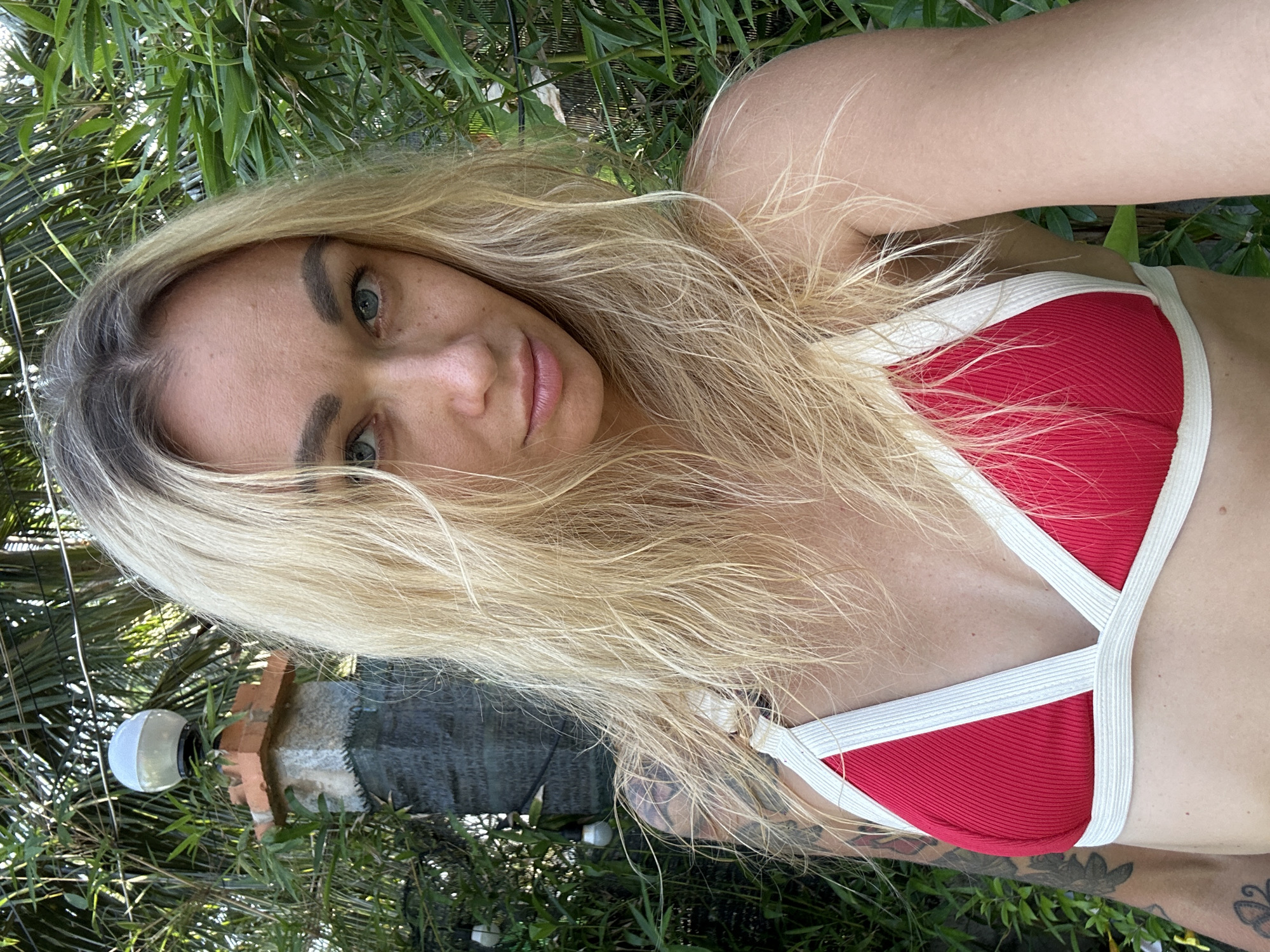 A woman with blonde hair and blue eyes is taking a selfie outdoors. She is wearing a red bikini top with white trim. She has tattoos on her left arm. Behind her, there is lush green foliage, including palm trees. In the background, a stone pillar topped with a spherical light fixture is visible. The scene appears to be in a tropical environment, potentially in Mũi Né, Vietnam, known for its beaches and palm trees.