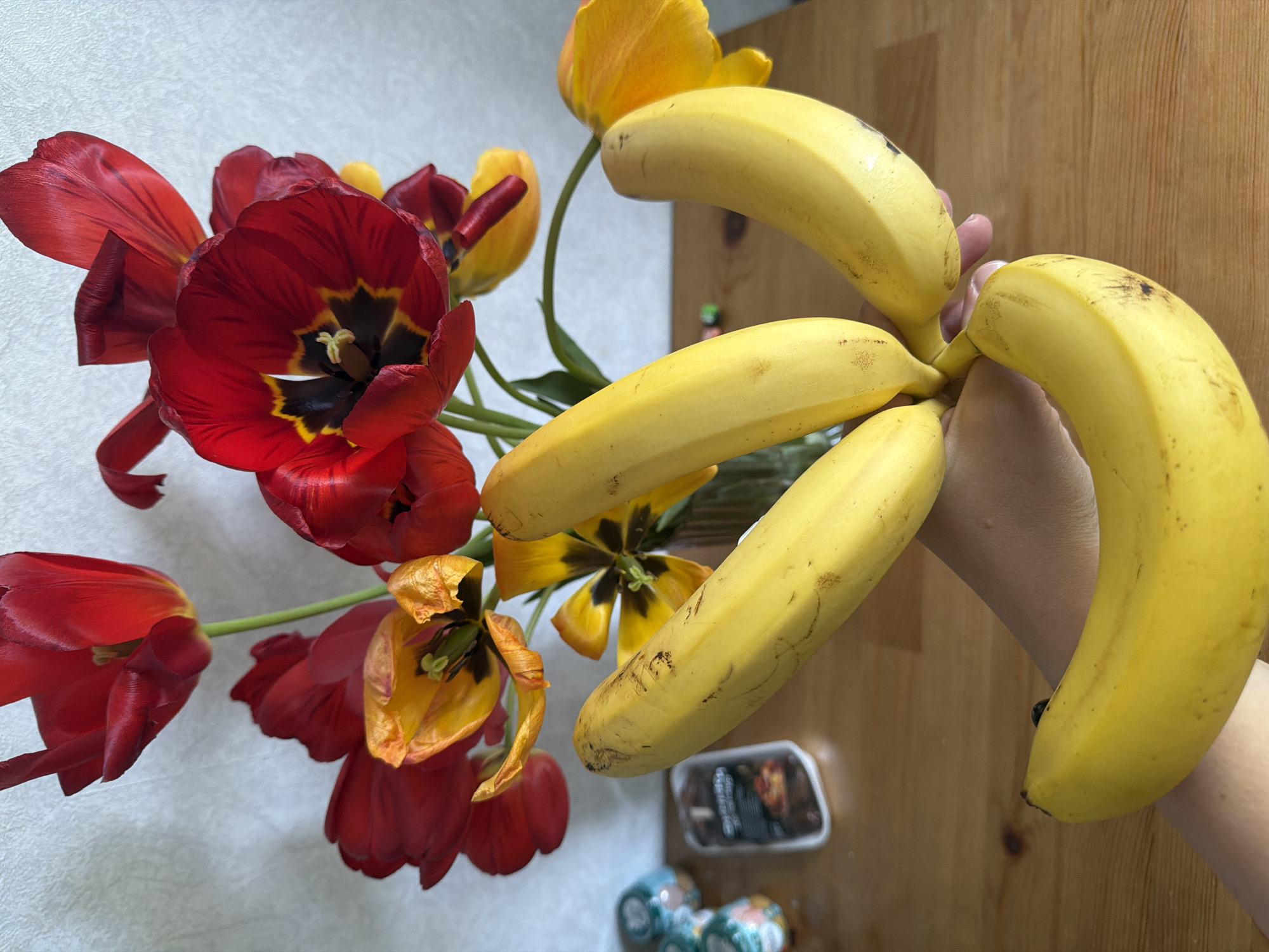A high-angle shot shows a bunch of yellow bananas being held in a person's hand, positioned to the right of a bouquet of red and yellow tulips in various stages of bloom. The setting appears to be indoors. The background consists of a white surface with a slight texture to the left and a wooden surface to the right. Below the bananas and flowers, there is a small, rectangular container with visible contents, and two small, patterned objects of light blue color. There is no indication in the photo that it was taken in Novosergievka, Russia.