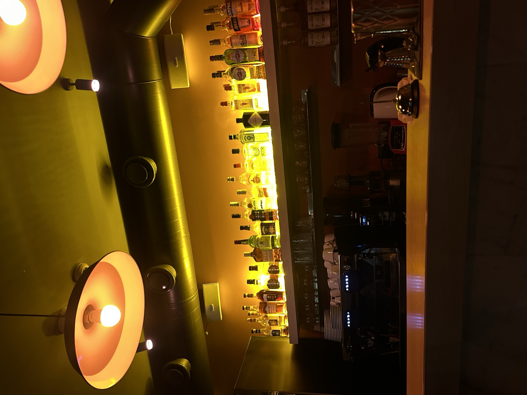 The image depicts the interior of a bar, possibly in Yerevan, Armenia, although this location is unconfirmed. The scene is dominated by a bar shelving unit displaying rows of liquor bottles backlit with bright yellow lighting. Adjacent to the liquor display is a countertop with bar equipment including blenders, serving glassware, and a commercial coffee machine. On the wall beside the shelving unit is a metal ventilation duct with circular vents, interspersed with rectangular fixtures. Two large pendant lamps with a warm orange glow are suspended from the ceiling, illuminating the wall. The overall lighting is low, creating a warm atmosphere within the space.