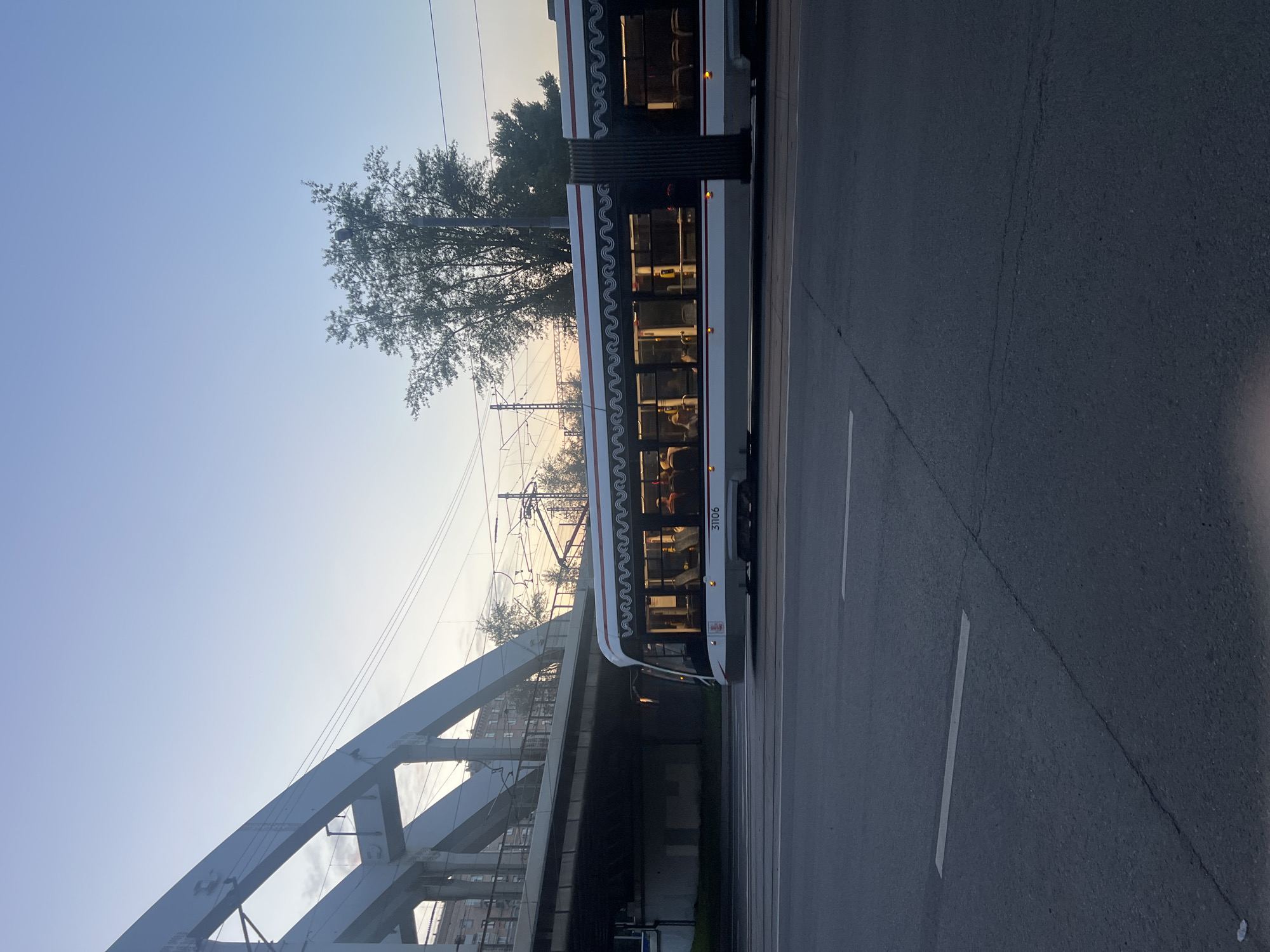 A side view of a white and red tram, number 31106, is captured in motion on a city street in Moscow, Russia, during a hazy sunset. The tram, with its windows illuminated from within, shows several passengers seated inside. The tram travels on tracks that run parallel to the road, which is marked with white lane lines. An elevated railway bridge, made of white girders and concrete supports, spans overhead. Beyond the bridge, trees and buildings are visible, and overhead power lines for the tram are strung across the sky. The sky is a pale blue with hints of pink and orange from the setting sun. The overall scene depicts a typical urban commute in Moscow.