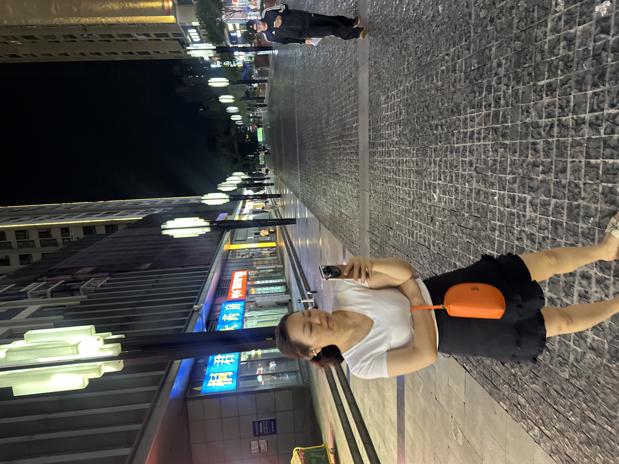 An eye-level shot captures a woman in the foreground on a cobblestone street at night, holding a mobile phone in her right hand and looking down at it. She is wearing a white t-shirt, a black skirt, and an orange clutch purse. Behind her, the street is lined with modern buildings, illuminated by decorative streetlights, suggesting an urban commercial area. Further down the street, another person in athletic wear is walking away from the camera, also looking at their phone. The scene appears to be in the city of Chongqing High-tech Zone, China.
