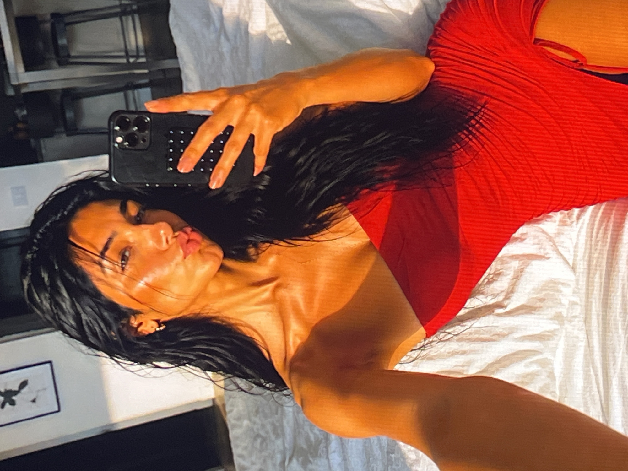 A medium shot shows a woman with long, dark, wet hair lying on a white, textured surface, possibly a bed. She is wearing a strapless red dress. The woman is taking a selfie with a black phone in her right hand, which has a studded case. She is pouting her lips and her face is illuminated by warm, golden light, suggesting it is either sunrise or sunset. In the background, out of focus, are kitchen counter stools and a framed picture on the wall. The lighting creates strong shadows and highlights on her skin, emphasizing her facial features and collarbone.
