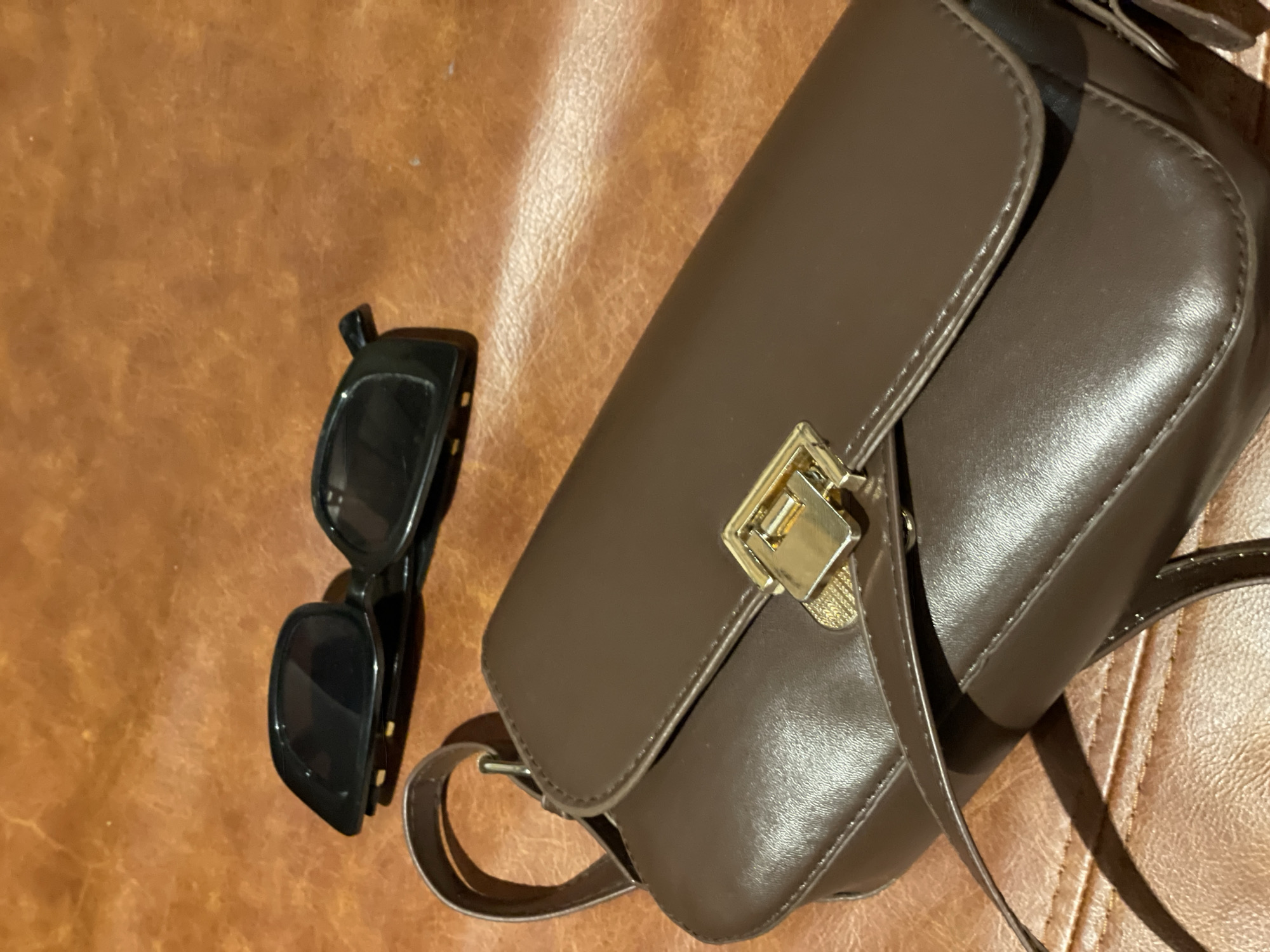 A brown leather purse with a gold clasp sits on a brown leather surface, possibly a table or a couch. Black sunglasses are placed behind the purse, angled slightly. The lighting suggests an indoor setting. There are no people or animals visible in the image. The background is out of focus, emphasizing the purse and sunglasses. There is no indication of the location being in Krasnoyarsk, Russia.