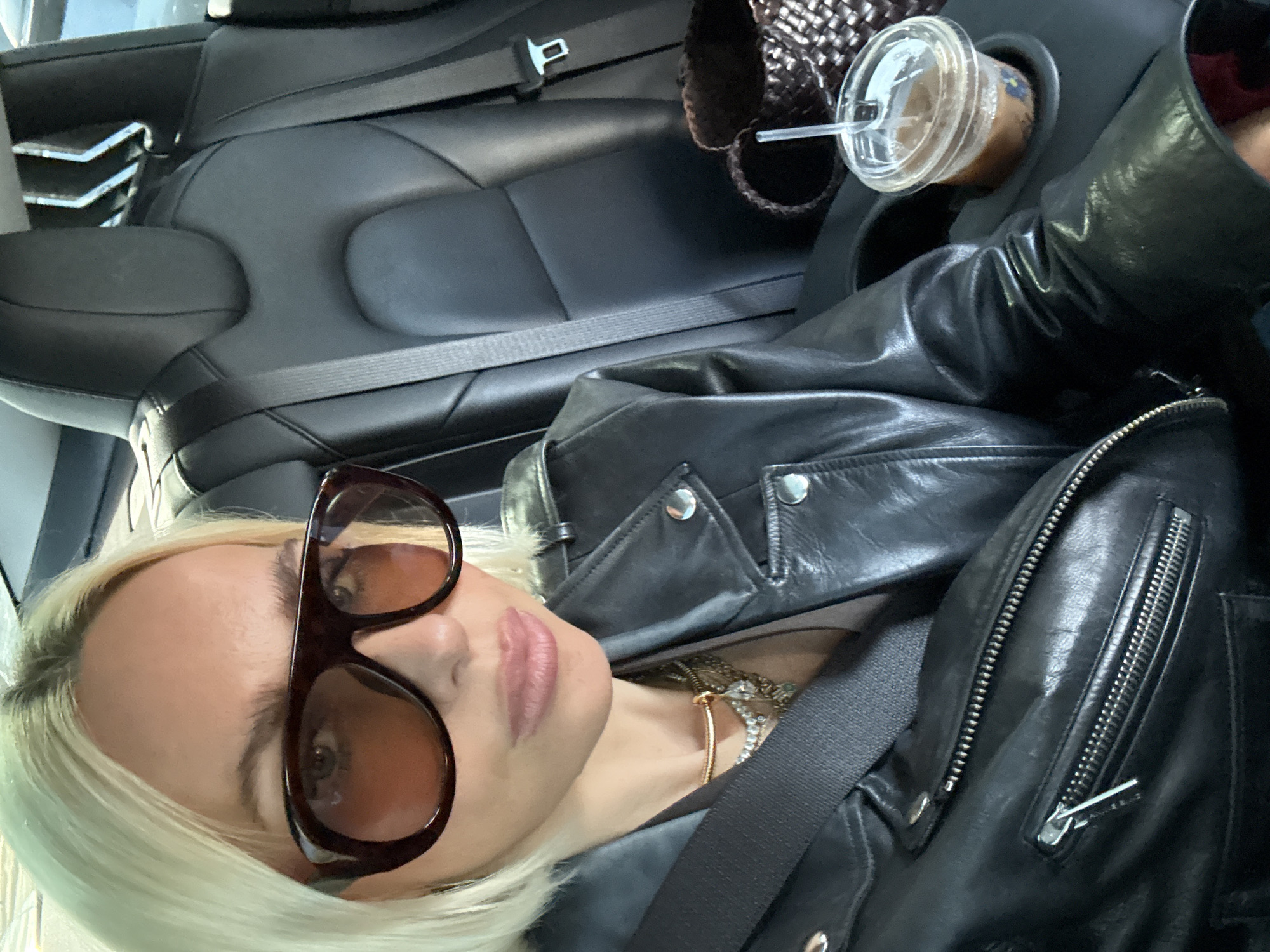 A person wearing a black leather jacket and large brown sunglasses sits in the backseat of a car with black leather seats. The person is wearing a seatbelt. A drink with a straw is in the cup holder. A brown woven bag is beside the drink. The photo was taken in the interior of the vehicle. It is not possible to determine the location within the city of Los Angeles without further context.