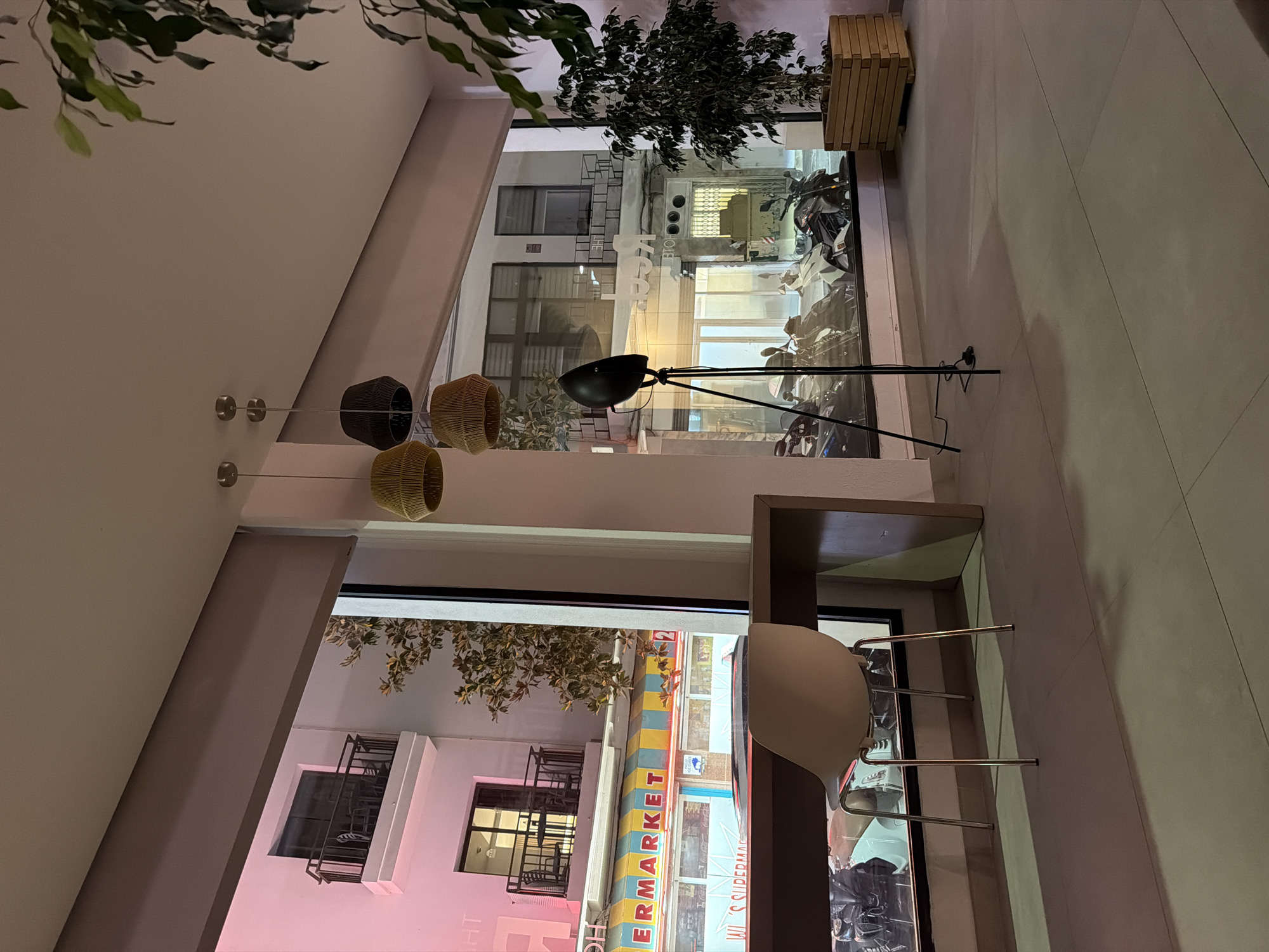 The image depicts an indoor scene with a large window offering a view of a street at night. Inside, a modern interior design is evident with a sleek black floor lamp and a white chair with a metallic frame. Three woven pendant lights hang from the ceiling, adding a touch of natural texture. To the right, a wooden planter holds a lush green plant.
The window reveals a street scene in Sant Antoni De Portmany, Spain. Buildings with balconies and windows are visible, illuminated by artificial lights. A sign for "SUPERMARKET" with colorful stripes is prominently displayed on one of the buildings. Several motorcycles are parked along the street, suggesting a bustling urban environment. The overall atmosphere is calm and serene, with the interior lights creating a warm glow, contrasting with the darker street outside. The time of day is clearly night, and no specific weather conditions are discernible. The image captures a moment of quiet observation from indoors, looking out at the vibrant street life.
