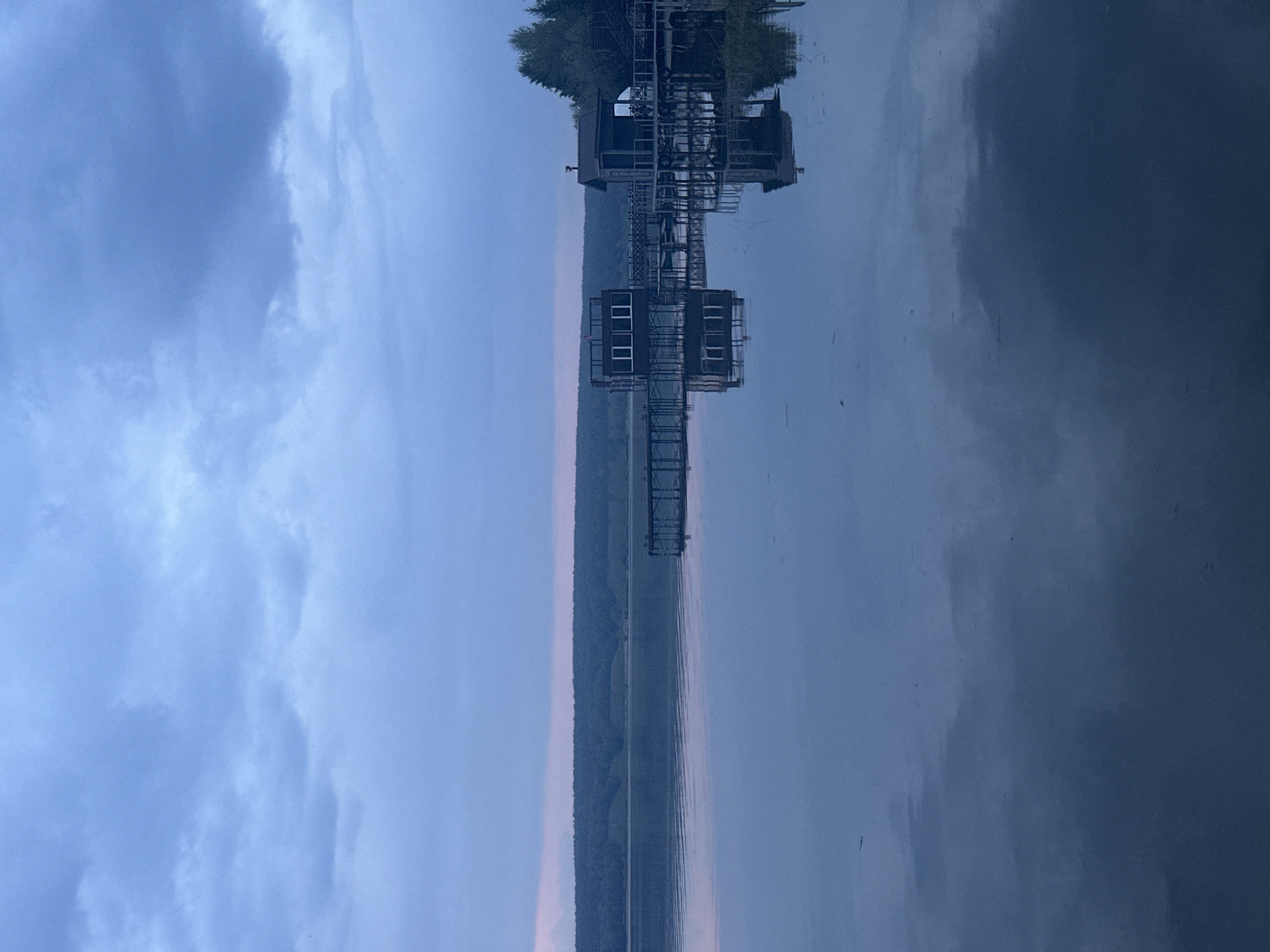 The image depicts an upside-down reflection in water of a structure resembling a boat lift or a pier with buildings on it. The sky is visible with cloud cover, suggesting it might be twilight or dusk. A line of trees or forest borders the water's edge. The location is specified as Aviator, Russia, but no landmarks or specific identifying features confirm this. The buildings appear to be utilitarian or industrial.