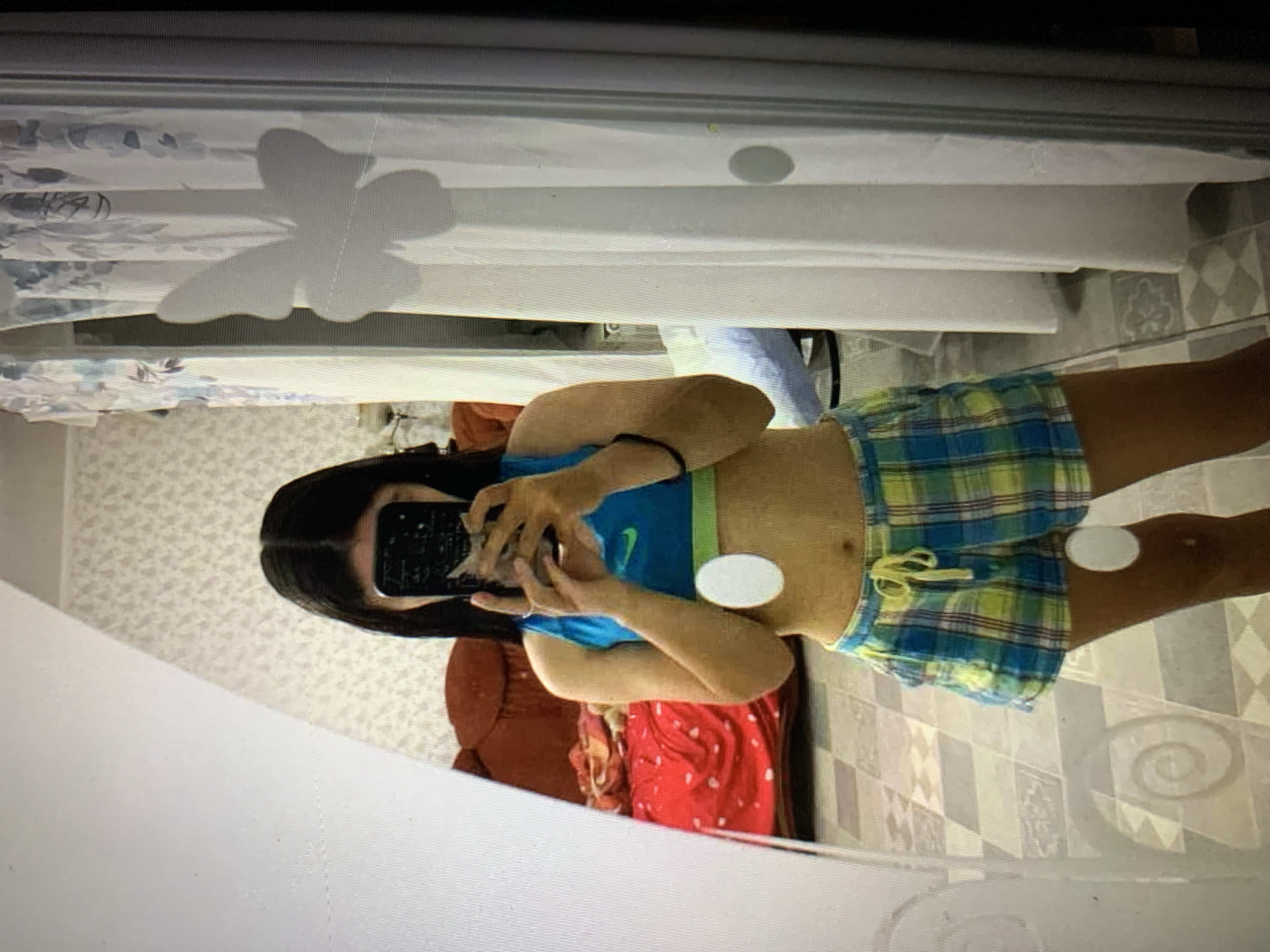 A young female with long, dark hair is standing in front of a mirror, taking a selfie. She is wearing a blue sports bra and plaid shorts. She is holding a black smartphone in her hands to capture the reflection. The background shows a room with a red patterned bedspread on a couch, a white patterned wallpaper, and a white curtain with a butterfly decal. The flooring appears to be tiled. The scene is consistent with a residential interior, potentially in Lymanka, Ukraine, given the context.