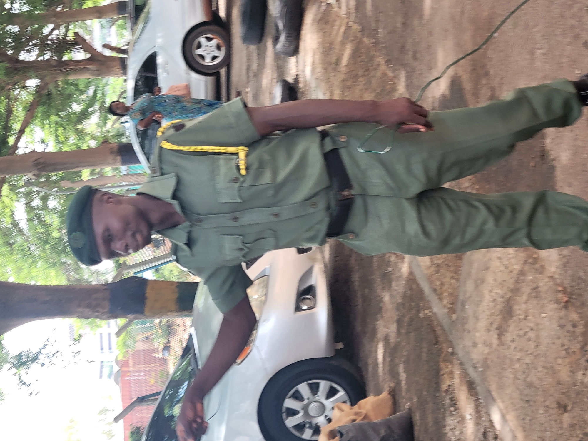 A man in an olive green uniform, wearing a matching cap, stands next to a silver car. He is holding a thin, green wire in his right hand. He has a black belt, and a yellow rope is draped over his left shoulder.
A second silver car is visible in the background, with a woman seated inside. Trees line the background. The ground is paved and sandy.
The location appears to be in Abuja, Nigeria, based on the uniform and the general environment.
