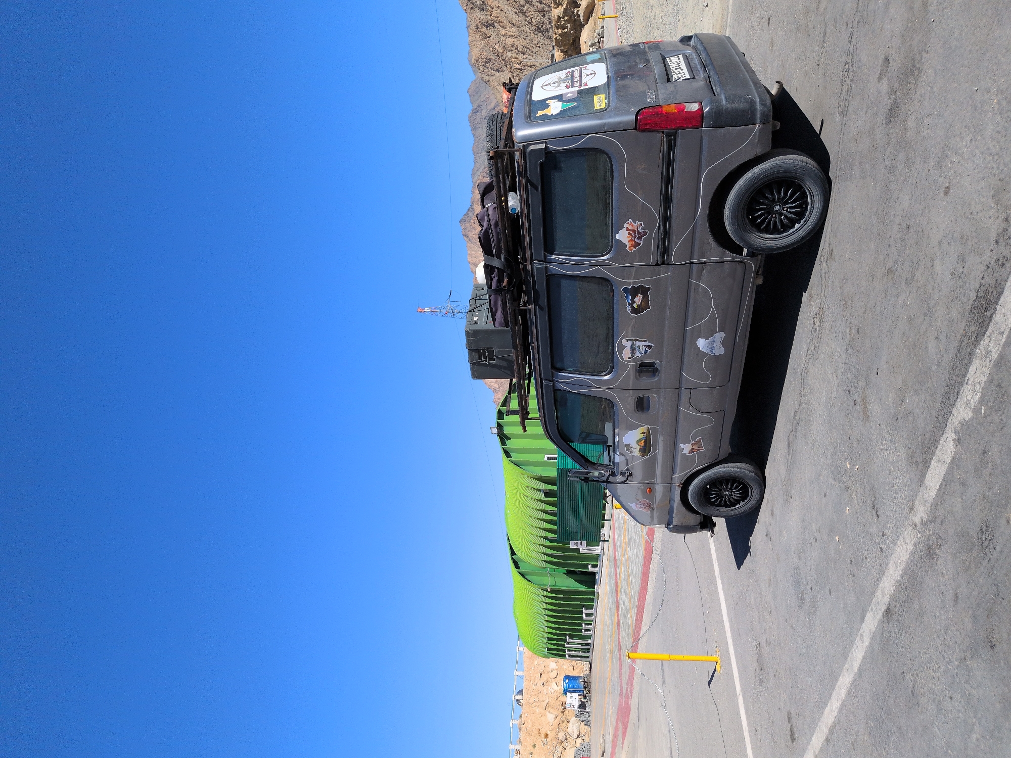 The image shows a gray van parked in an outdoor area with a clear blue sky. The van is covered in stickers depicting maps, scenic views, and various cartoon characters. It has a roof rack loaded with luggage, including a black suitcase, a spare tire, and what appears to be a rolled-up mat. The van is positioned on a paved surface with white and red-lined parking demarcations. In the background, there are two green, arched structures that resemble large tents or covered shelters. To the left of these structures, there are some blue containers and a yellow bollard. The landscape behind the van and structures consists of arid, rocky hills. The scene appears to be a rest stop or service area, possibly along a travel route. The license plate on the back of the van is partially visible, reading "SL3BNWAD22". There are no people visible in the image. The location is identified as Chumkar, India.