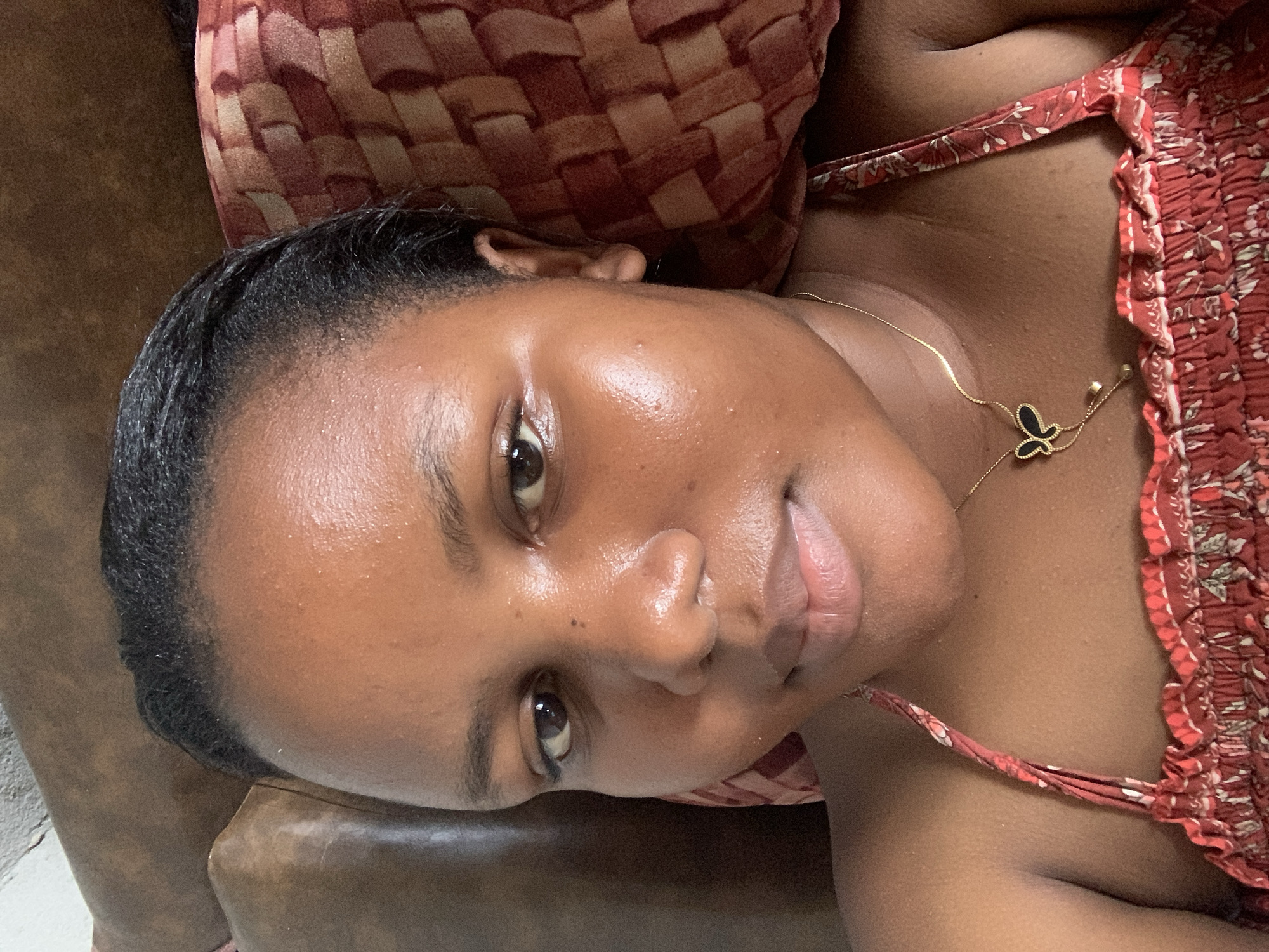 A close-up, angled shot of a woman lying down. She has dark hair styled back from her forehead, dark eyebrows, and dark eyes. Her skin is a medium brown tone. She is wearing a red and white patterned top with spaghetti straps, and a delicate gold necklace with a butterfly pendant. She appears to be resting her head on a brown and red patterned pillow or cushion, which is itself on a brown surface, possibly a couch or chair. There is no visual evidence to indicate a location in Dar es-Salaam, Tanzania.