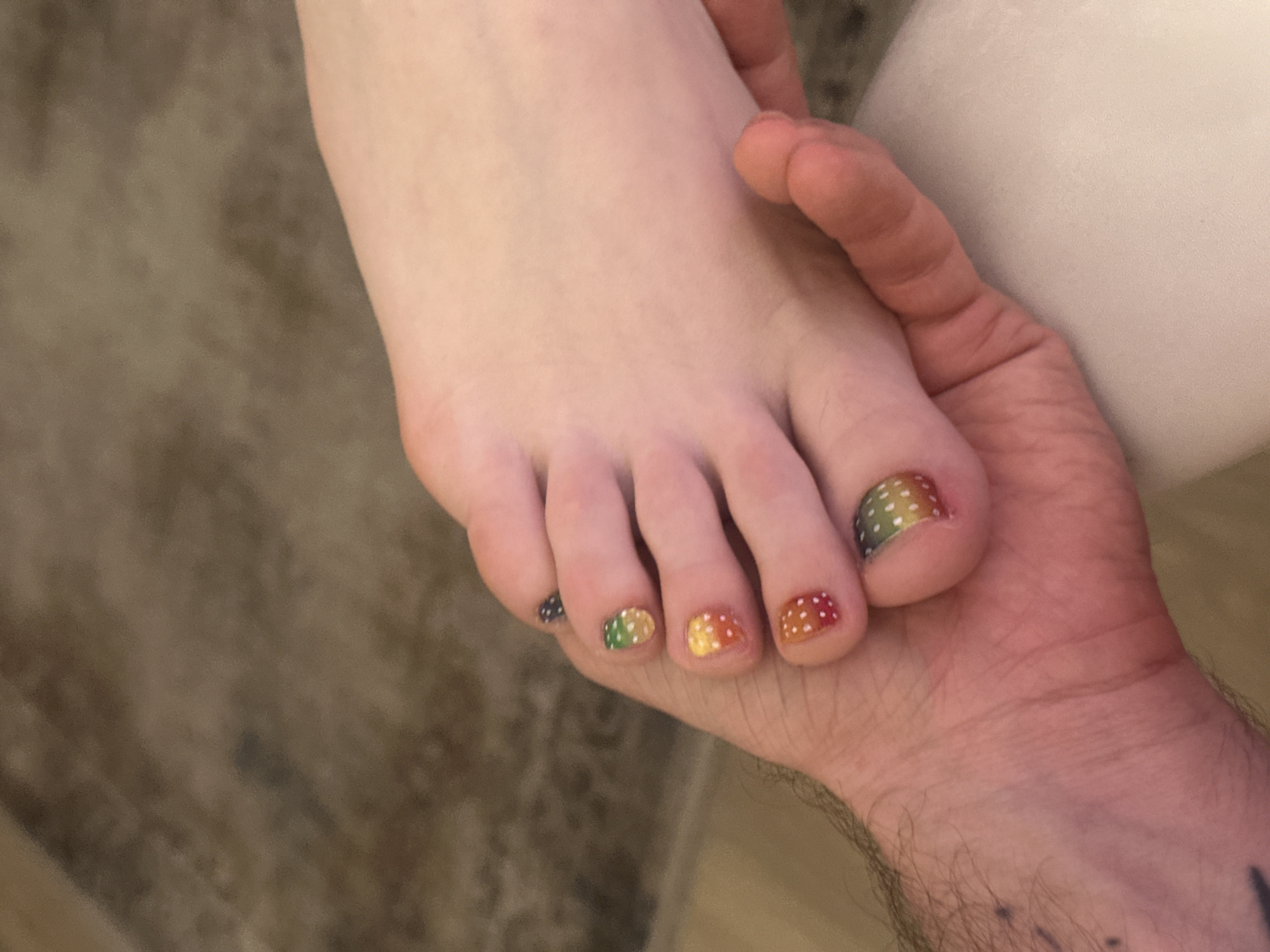 A close-up shot depicts a fair-skinned foot with painted toenails resting in a person's hand. The toenails are painted with a rainbow gradient effect, each decorated with small white dots. The hand holding the foot appears to belong to a different person, characterized by a slightly darker skin tone and visible hair on the wrist. The backdrop includes a neutral-toned rug or carpet. The photograph lacks any information that can point to Moscow, Russia.
