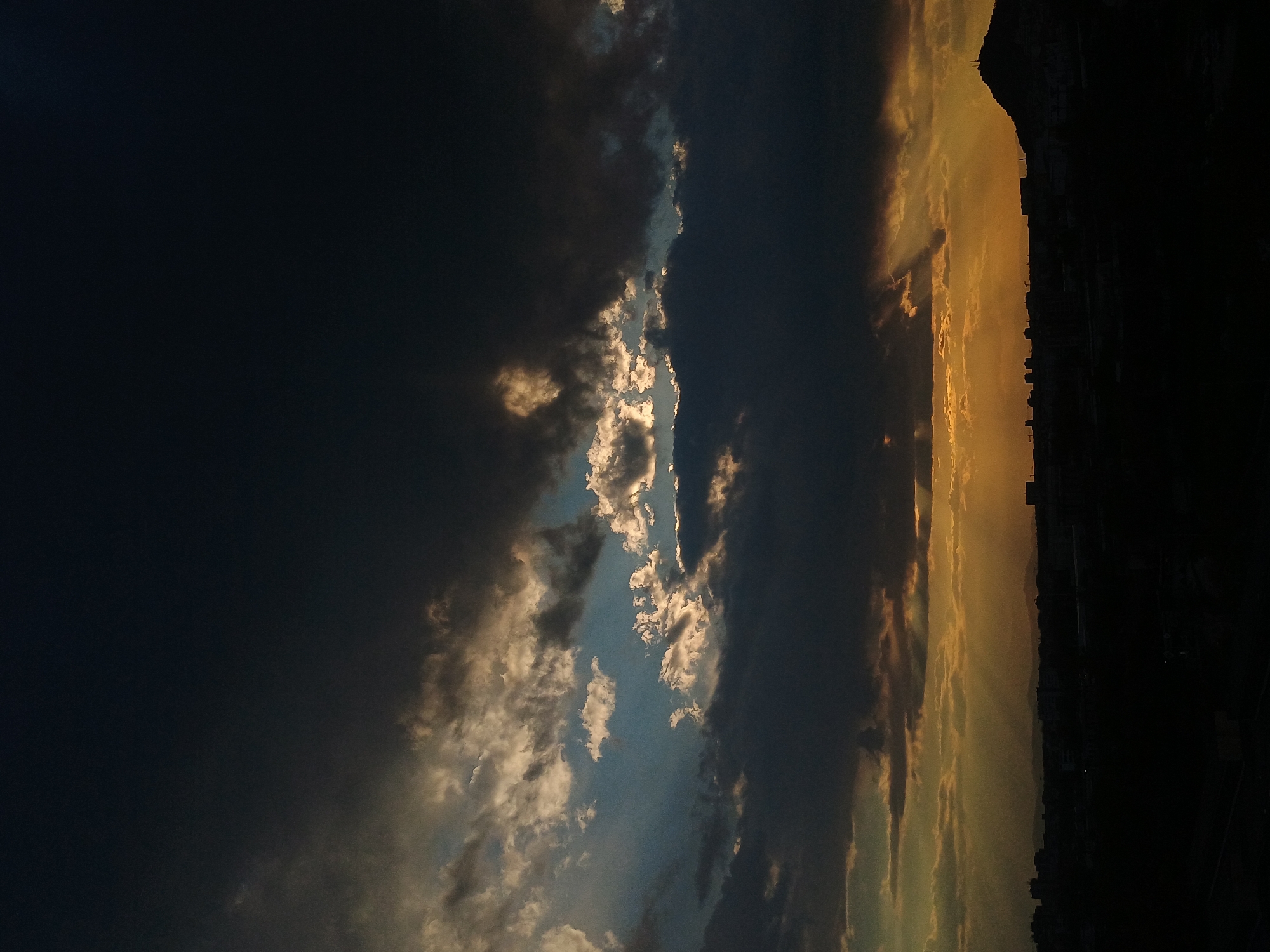 The image shows a scene in Plovdiv, Bulgaria, possibly at dawn or dusk. The sky is a mix of dark, ominous clouds and lighter clouds reflecting sunlight. On the right, a silhouette of the city skyline is visible against the bright sky, with buildings and possibly hills discernible. The overall impression is one of dramatic weather over an urban landscape.